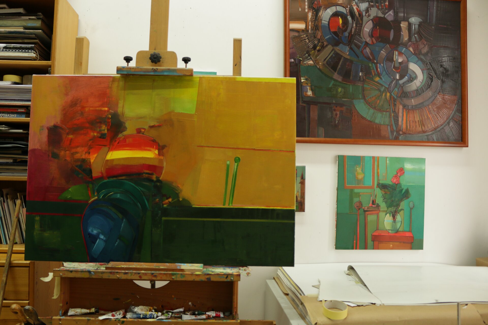 Ilustracja przedstawia fragment wnętrza pracowni malarskiej. W centrum kadru znajduje się sztaluga z abstrakcyjnym obrazem olejnym w żółto‑czerwono‑zielonej tonacji. Za nim ustawiony jest wysoki regał z książkami. Po prawej stronie stoi niska, biała szafka z leżącą na niej szarą teczką z rysunkami. Na białych ścianach pomieszczenia wiszą kolorowe obrazy.