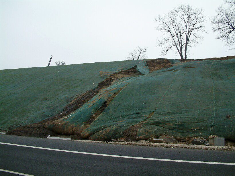 Na zdjęciu jest osuwające się zbocze pagórka. U jego podstawy biegnie asfaltowa droga. Ziemia na zboczu jest porozsuwana, widoczne są szczeliny i doły. Zbocze pokryte jest zieloną tkaniną, która zabezpiecza przed dalszymi obsunięciami ziemi na drogę. 