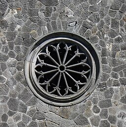 Zdjęcie pokazuje okrągłe okno w murze zwane rozetą. Wnętrze okna wypełnia ożebrowanie - żebra ułożone są od środka okna promieniście, tworząc płatki kwiatu.  