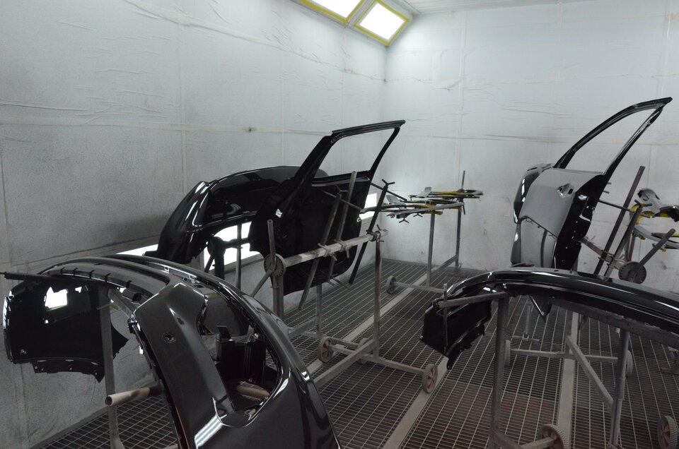 Ilustracja przedstawia komorę lakierniczą. Na stelażach zamieszczony zostały elementy karoserii, drzwi, błotniki, które zostały już polakierowane na kolor czarny. Komora ma białe ściany i podłogę z kratownicy.