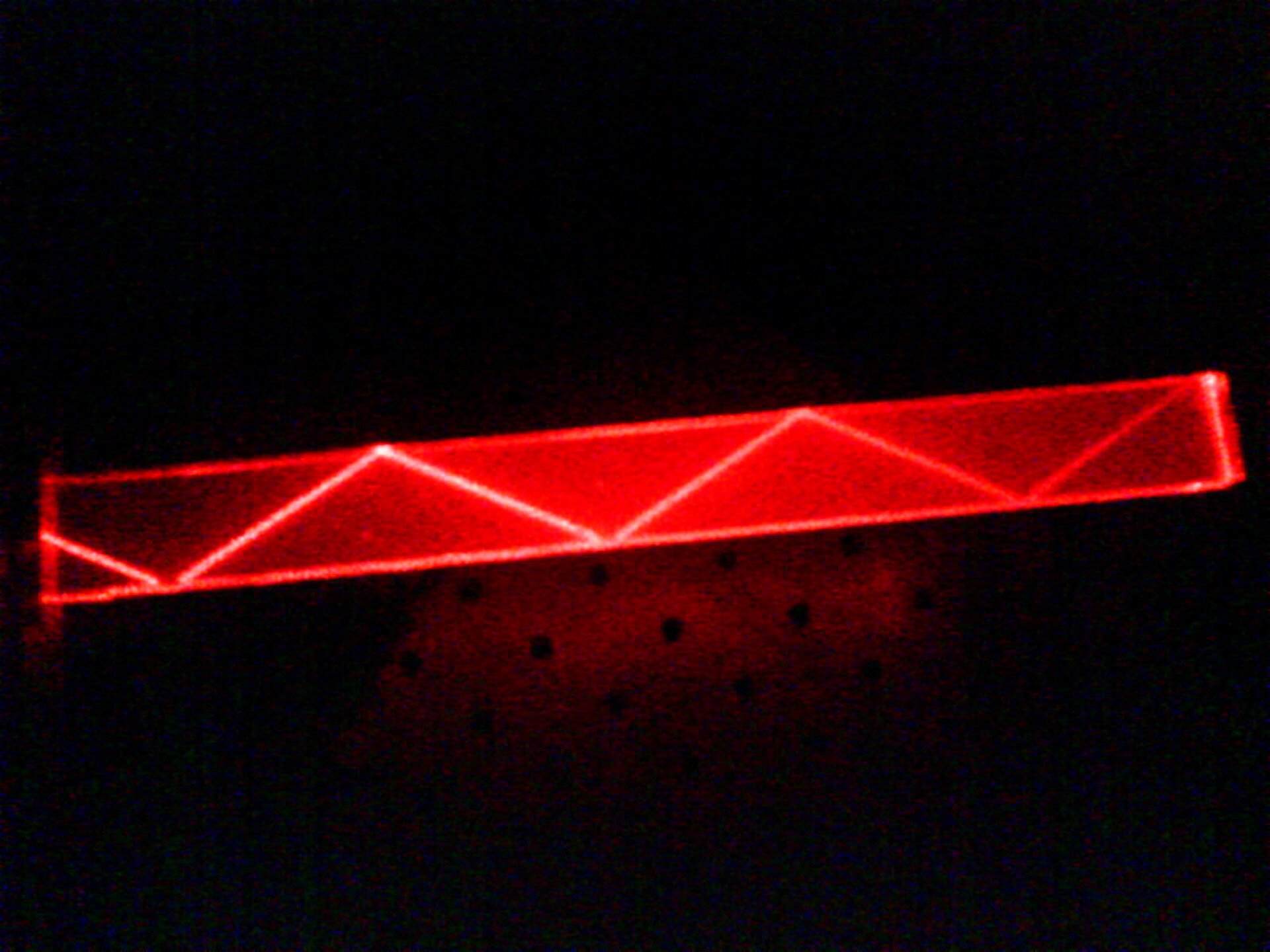 Zdjęcie przedstawia całkowite wewnętrzne odbicie. Na czarnym tle widać przezroczysty element w kształcie prostopadłościanu. Wewnątrz widać czerwone nieco rozmazane odcinki tworzące zygzakowatą linię. Światło laserowe wielokrotnie odbija się od wewnętrznych ścianek przezroczystego materiału.