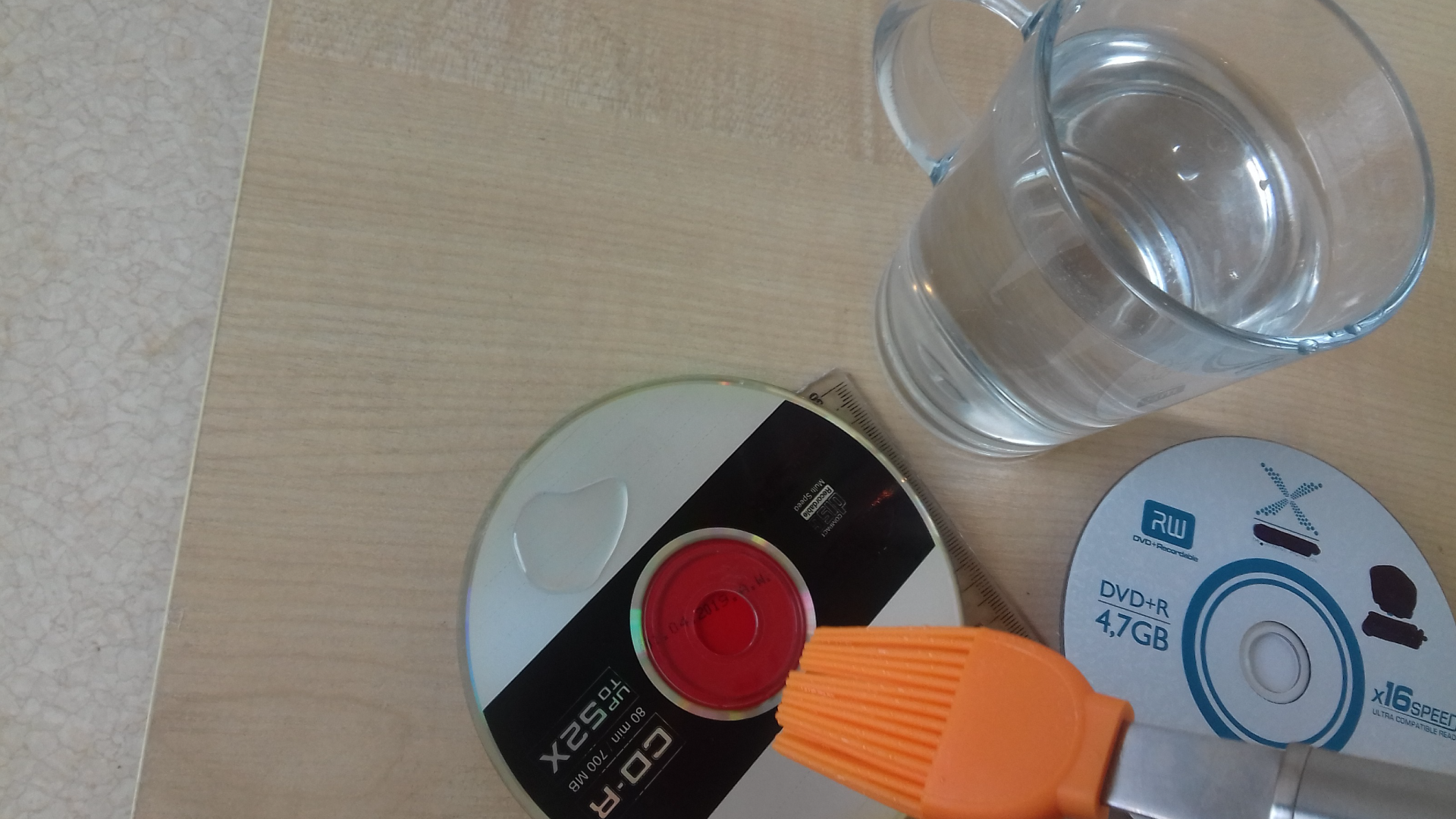 Zdjęcie przedstawia przedstawia 2 dyski optyczne, linijkę oraz szklankę wody i pędzel nad jedną z płyt. Na płycie widoczna jest woda.