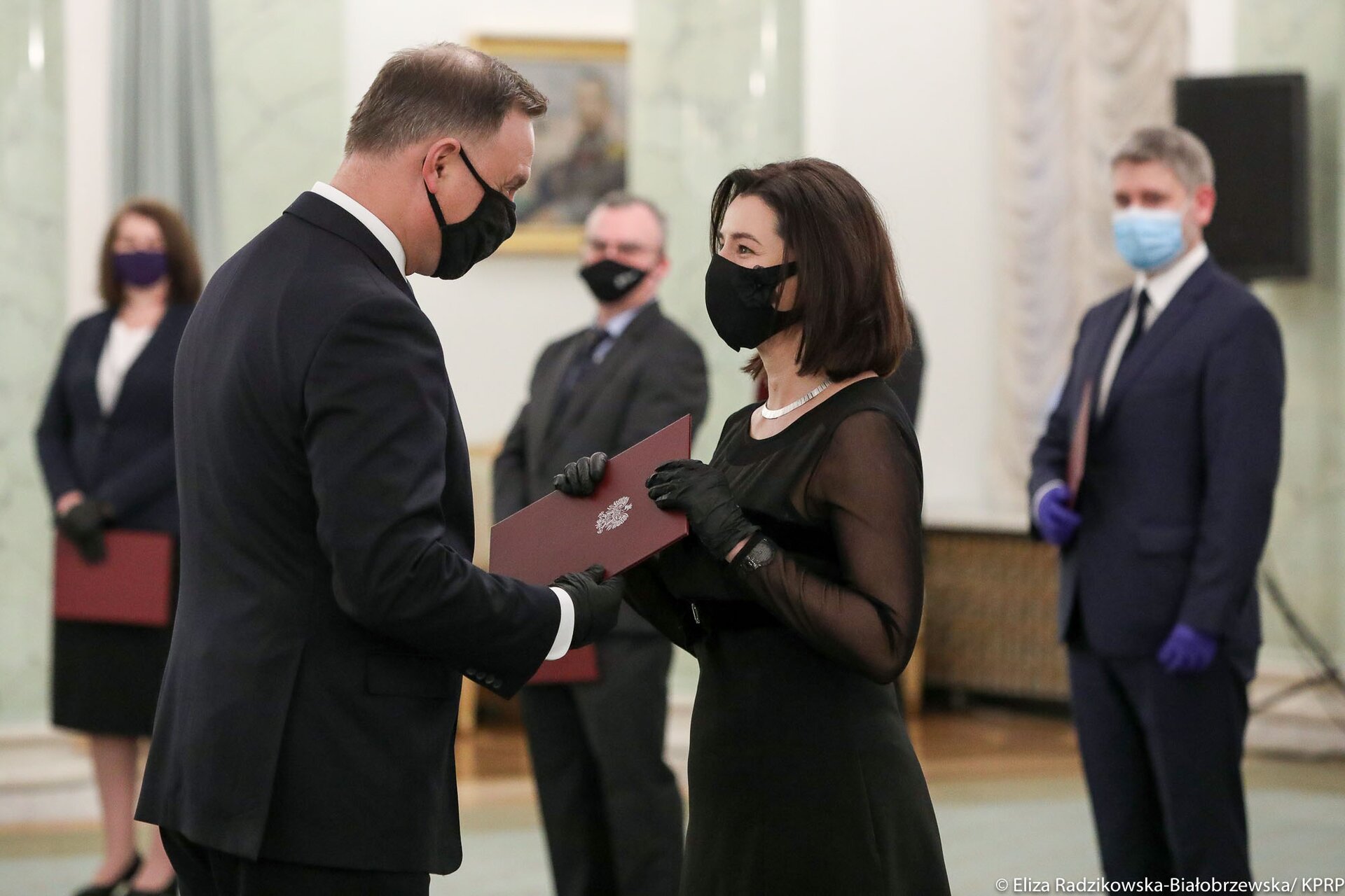 Kliknij żeby powiększyć Zdjęcie, które przedstawia wręczenie nominacji sędziowskich przez Prezydenta RP Andrzeja Dudę. Uroczystość jest w Pałacu Prezydenckim. Nominacje mają miejsce w czasie trwania obostrzeń epidemiologicznych - Prezydent RP i nominowani sędziowie mają ubrane maseczki i założone rękawiczki. Prezydent RP wręcza akt powołania w czerwonej teczce. Mężczyźni ubrani są w czarne lub granatowe garnitury. Jedna z kobiet, która odbiera akt z rąk Prezydenta RP ma ubraną czarną sukienkę. Natomiast kobieta, która odebrała akt nominacji i z innymi powołanymi już do pełnienia urzędu stoi w szeregu i jest ubrana w granatową garsonkę. Po lewej stronie na ścianie wisi obraz. Za stojącymi sędziami stoi głośnik.