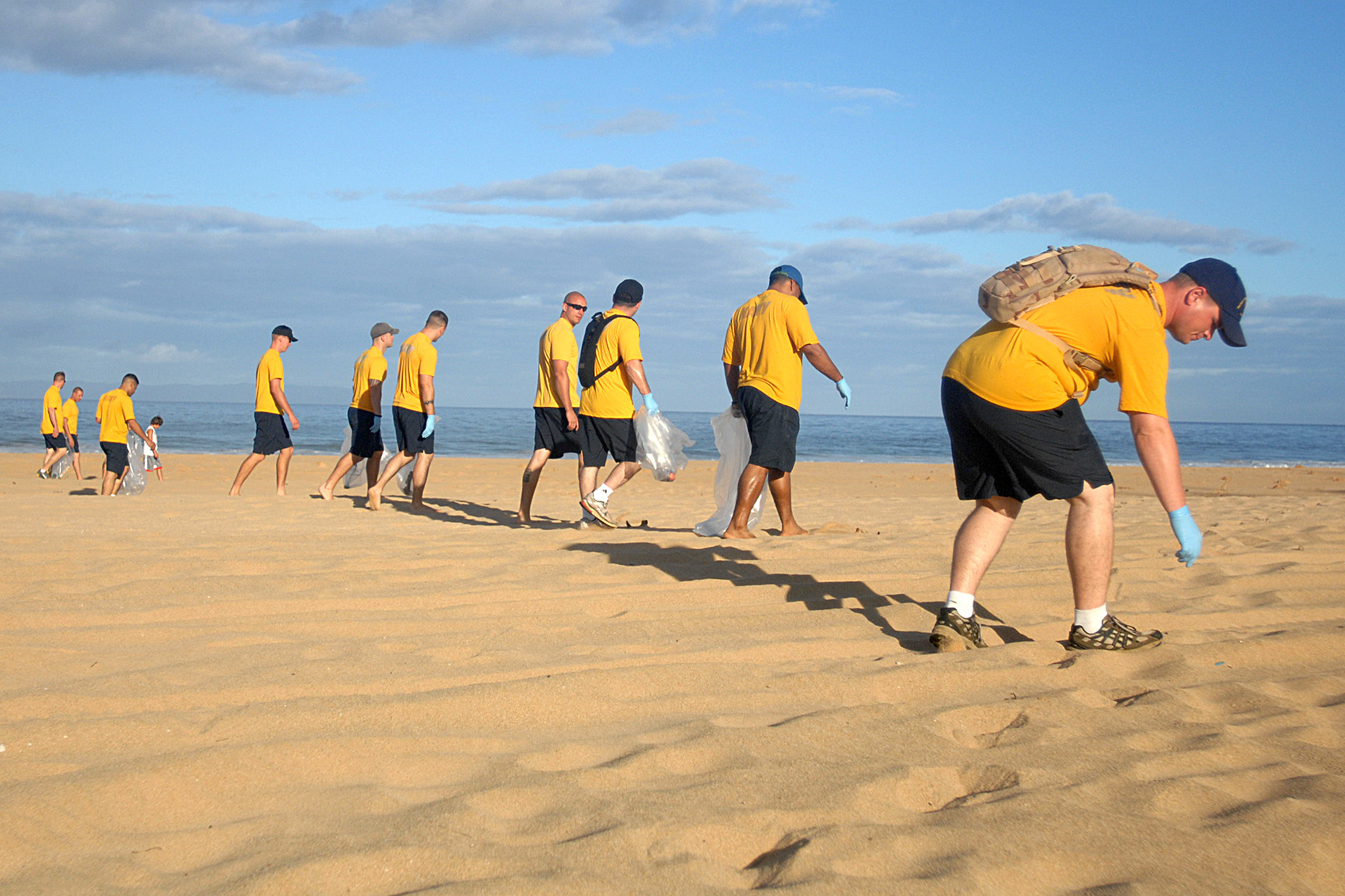 Fotografia przedstawia grupę osób znajdujących się w jednej linii w żółtych koszulkach i czarnych spodniach, które zbierają śmieci z plaży. W tle morze i błękitne niebo. 