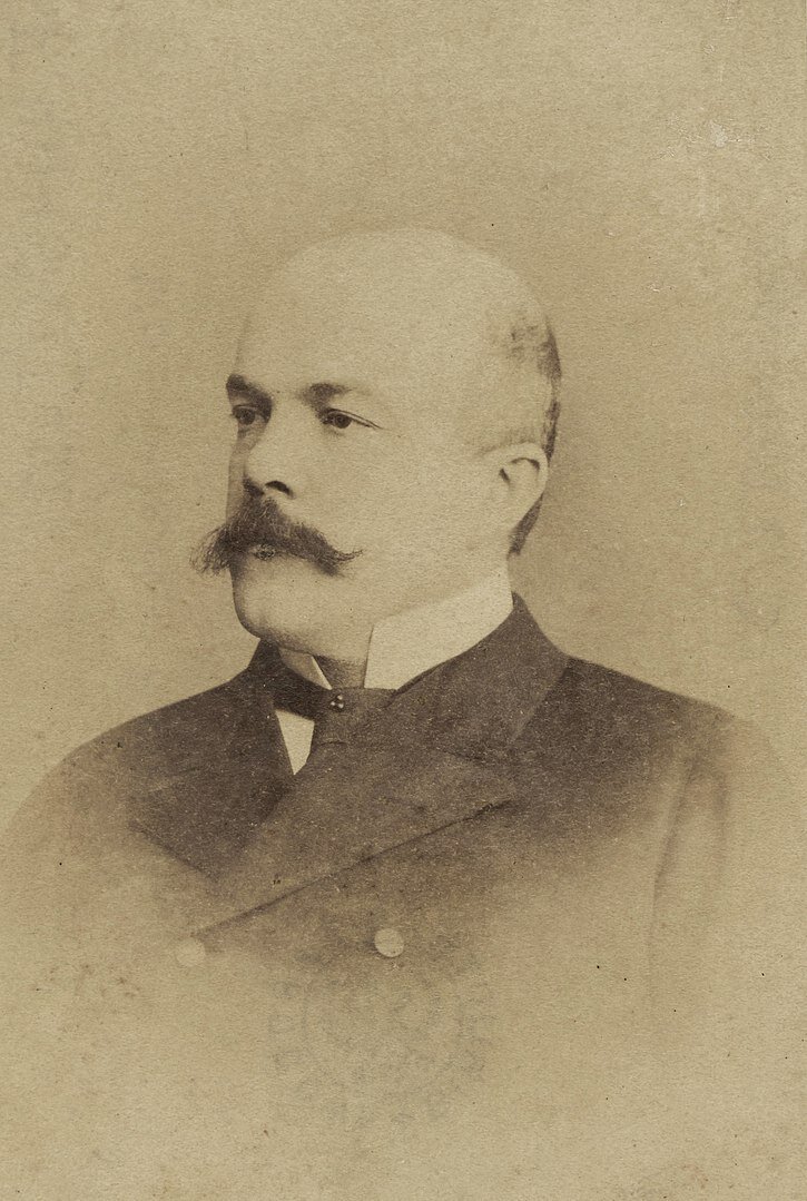 Zdjęcie przedstawia mężczyznę. Jest łysy, ma gęste wąsy. Ubrany jest w surdut.