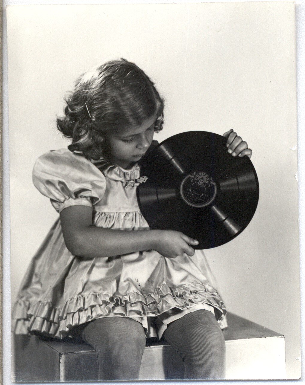 Na zdjęciu dziewczynka ubrana w zdobioną, jedwabną sukienkę i rajstopy reklamuje płytę z nagraniem "Bajek" A. Mickiewicza. Trzyma ona płytę w obu rękach i ma głowę zwróconą w tę stronę. Reklama pochodzi z 1935 roku.