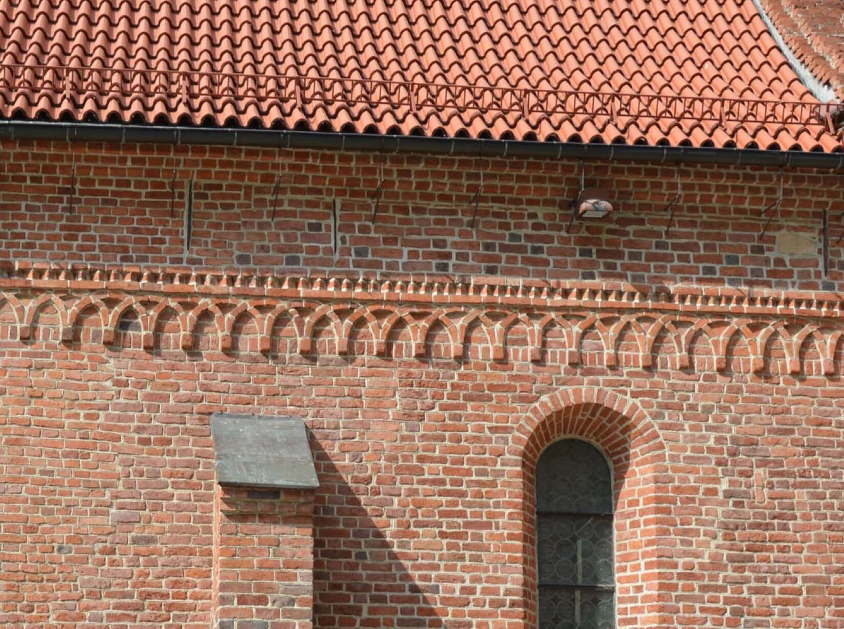 Fotografia przedstawia gzyms na fragmencie budynku zbudowanego z cegieł w odcieniach pomarańczy. Detal przedstawia zdobienie, które swoim kształtem przypomina łuki nad którymi biegnie balustrada.