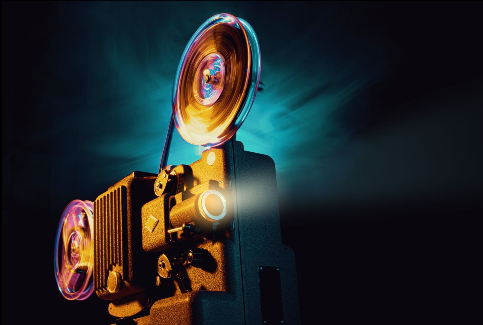 Zdjęcie przedstawia projektor filmowy dawnego typu, z założoną taśmą filmową na rolce. Projektor ukazany jest w ciemnościach podczas emisji filmu, na co wskazuje łuna światła wydobywająca się z aparatu. Delikatne oświetlenie ukazuje szczegóły budowy projektora. W pomieszczeniu zalega ciemność.