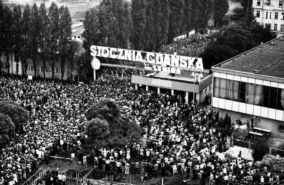 Czarno-biała fotografia przedstawiająca brama nr 2 Stoczni Gdańskiej im. Lenina i tłumy solidaryzujących się ze strajkującymi mieszkańców Gdańska (1980 r.). 