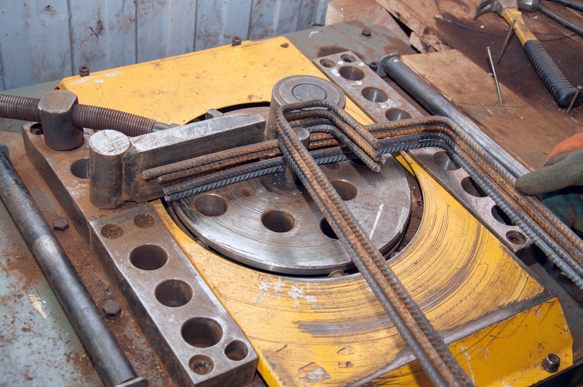 Zdjęcie przedstawia giętarkę mechaniczną o żółtej obudowie. W środku giętarki znajduje się kilka odginanych prętów.