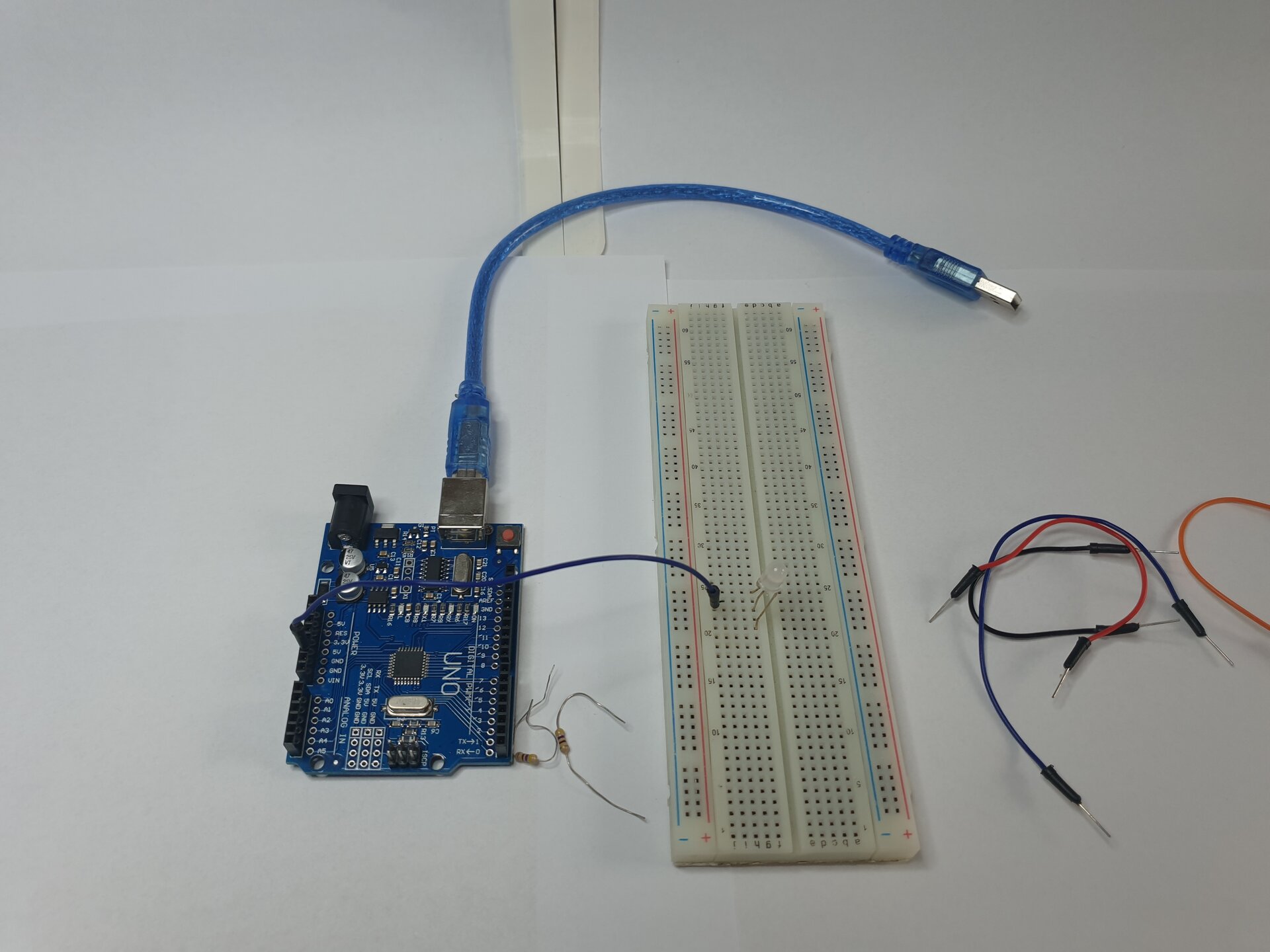 Zdjęcie przedstawia moduł Arduino z podłączonym do niego kablem USB, dwa rezystory, płytkę stykową, w którą wpięto diodę oraz leżące obok płytki przewody. Połączono moduł z płytką za pomocą przewodu wpiętego w module w 5 V i w płytce w tym samym wierszu, co środkowa nóżka diody.