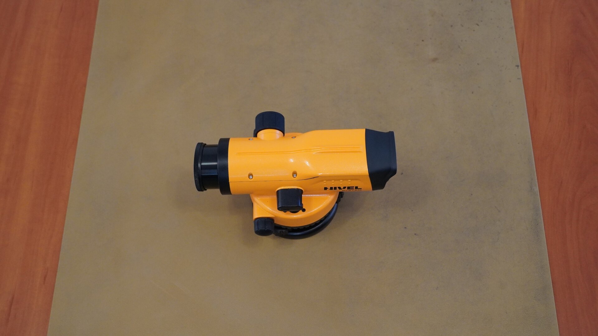 Zdjęcie przedstawia widok na niwelator z góry. Widać podłużną lunetę zakończoną okularem z jednej strony, a obiektywem z drugiej. Po bokach znajdują się czarne pokrętła. Luneta umieszczona jest na okrągłej podstawie.