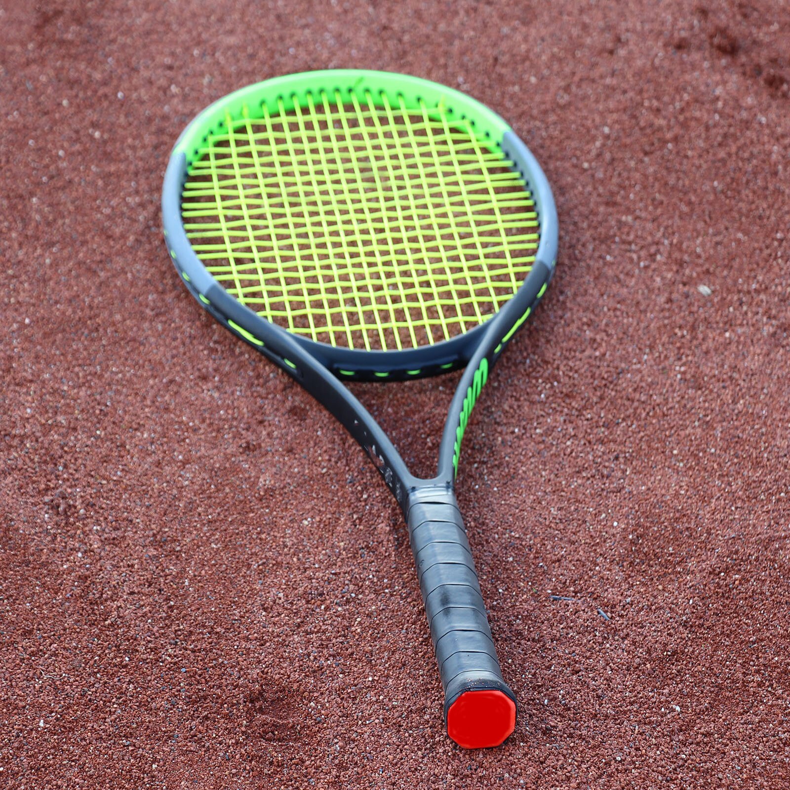 Ilustracja przedstawia rakietę do tenisa, leżącą na czerwonym, piaszczystym korcie. Rakieta ma żółty naciąg oraz zielono‑czarną ramę.