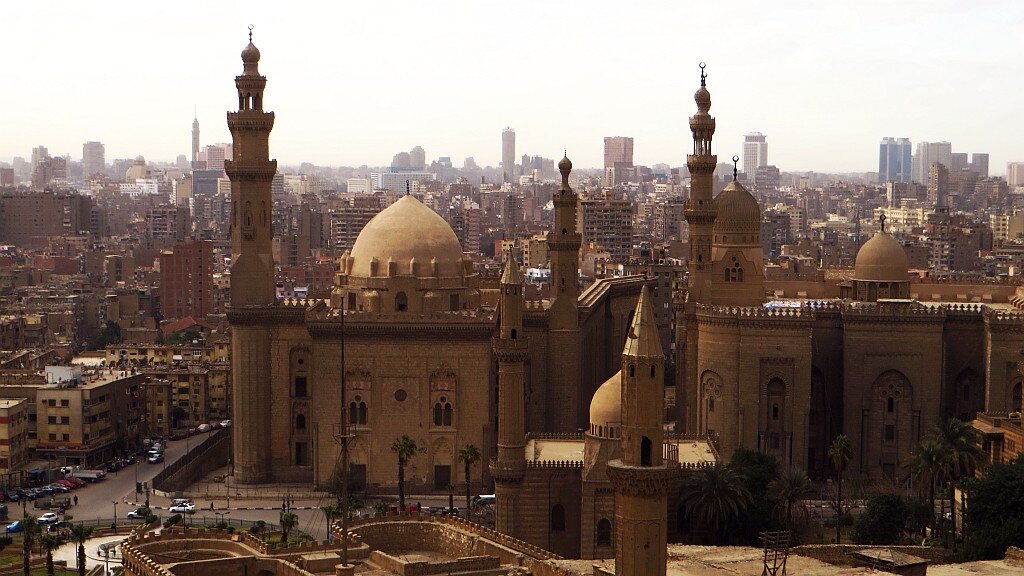 Zdjęcie przedstawia duże miasto o gęstej zabudowie. Na pierwszym planie są meczety - budynki z wieżami i kopułami. Za nimi stoją liczne wysokie i niskie budynki.