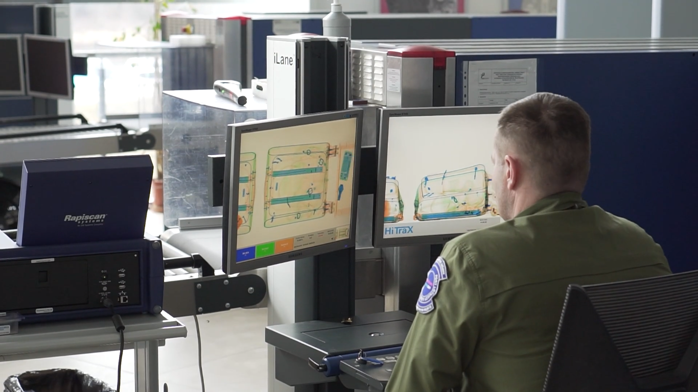 Zdjęcie przedstawia system wykrywania ładunków wybuchowych. Widoczny jest pracownik ochrony portu lotniczego siedzący przed dwoma monitorami. Na monitorach wyświetlane są prześwietlane walizki, w widoku z góry na lewym ekranie i w widoku z boku na prawym ekranie. Za monitorami znajduje się urządzenie z przenośnikiem taśmowym, na którym sprawdzane bagaże są umieszczane.
