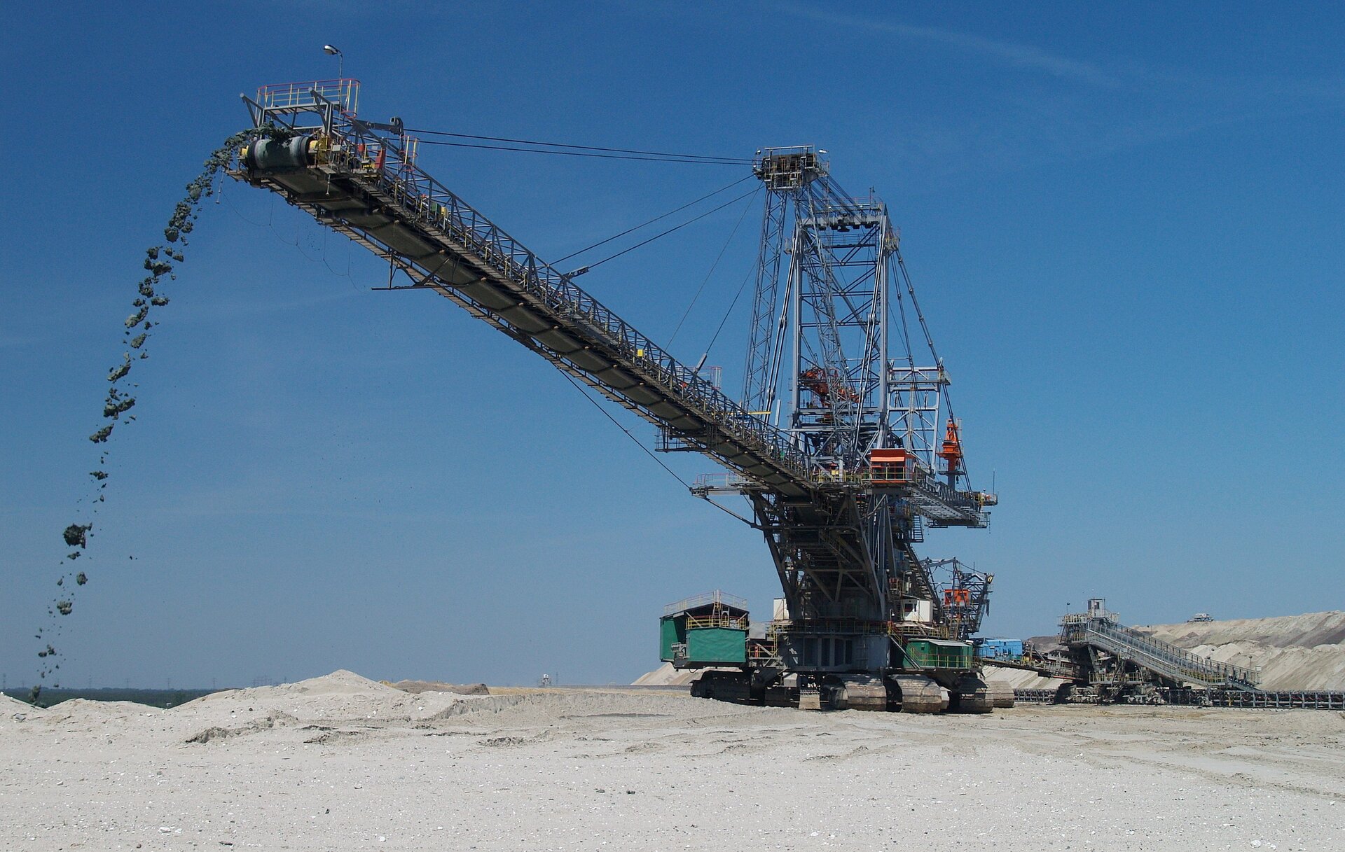 Zdjęcie przedstawia dużych rozmiarów maszynę stojącą na teren kopalni odkrywkowej. Podwozie składa się z kilku gąsienic umożliwiających ruch kołowy stalowej konstrukcji szkieletowej. Wysunięte długie ramię, na którym leży pas transmisyjny służący do przenoszeniu urobku. W górnej części widoczny system lin wzmacniających i stabilizujących konstrukcję