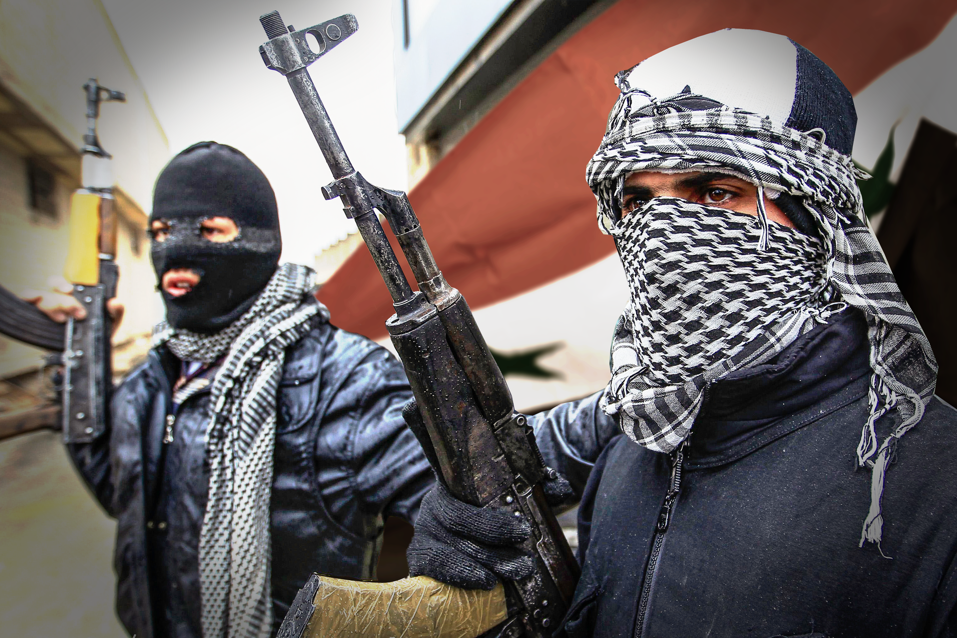 Kolorowe zdjęcie przedstawia dwóch mężczyzn stojących na ulicy. Mężczyźni mają zakryte twarze. Mężczyzna po lewej ma czarną kominiarkę. Mężczyzna po prawej białą czapkę i kraciastą chustę zawiązaną wokół twarzy. W prawej ręce każdy trzyma karabin. Karabin skierowany lufą w górę. W tle, za mężczyznami, flaga Syrii. Po lewej budynki mieszkalne.