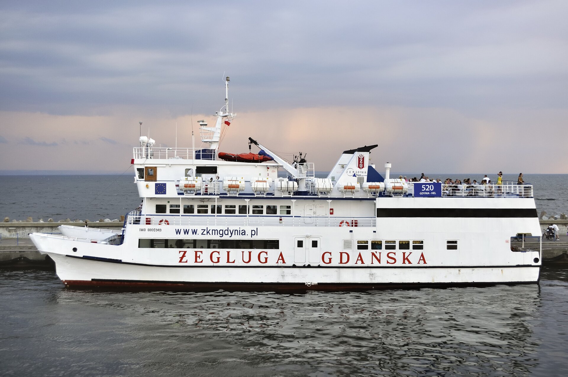 Zdjęcie przedstawiające niewielki prom na morzu. Ma kolor biały, na burcie napis czerwonymi literami „Żegluga Gdańska”. Wzdłuż burty linia okien, powyżej taras, na którym siedzą pasażerowie. Na górze promu anteny.