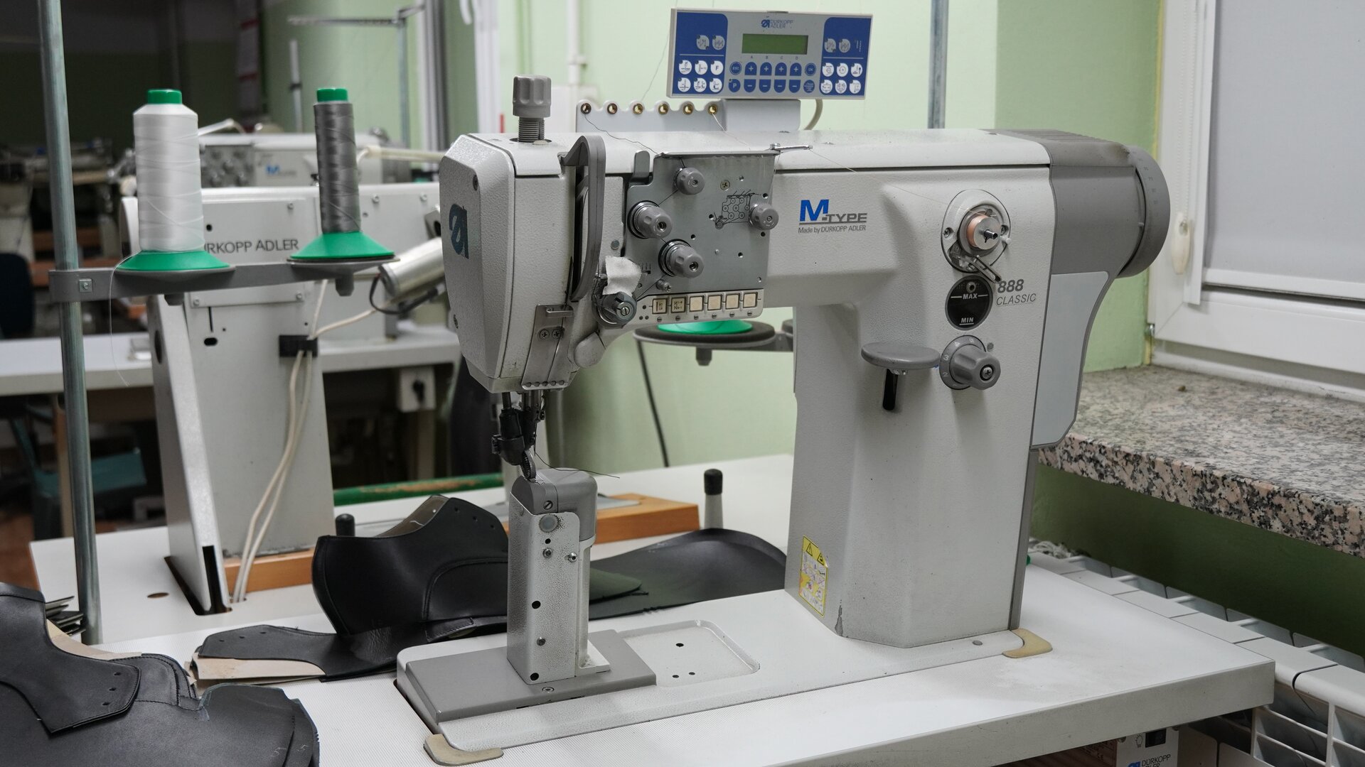 Zdjęcie przedstawia biało‑szarą maszynę słupkową do szycia, z przodu ma pokrętła, nad nią jest umieszczony panel sterowania. Stoi na blacie. Na blacie leżą zszyte cholewki. Nagranie tożsame z treścią.