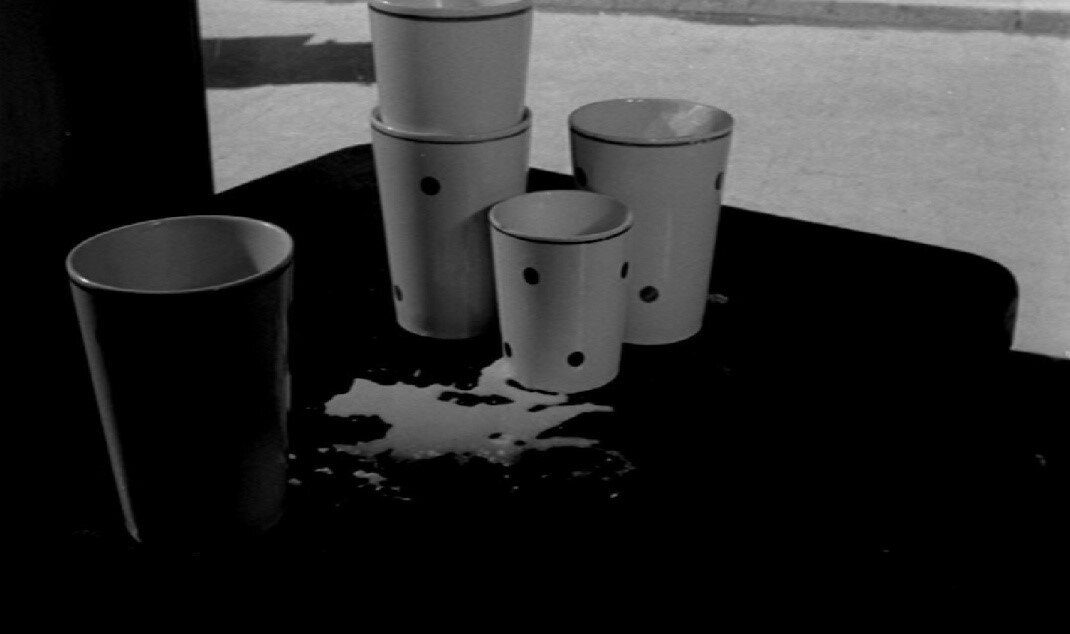Zdjęcie przedstawia stół, na którym stoi jeden duży kubek, trzy średnie i jeden mały kubek. Pomiędzy nimi jest rozlane mleko.