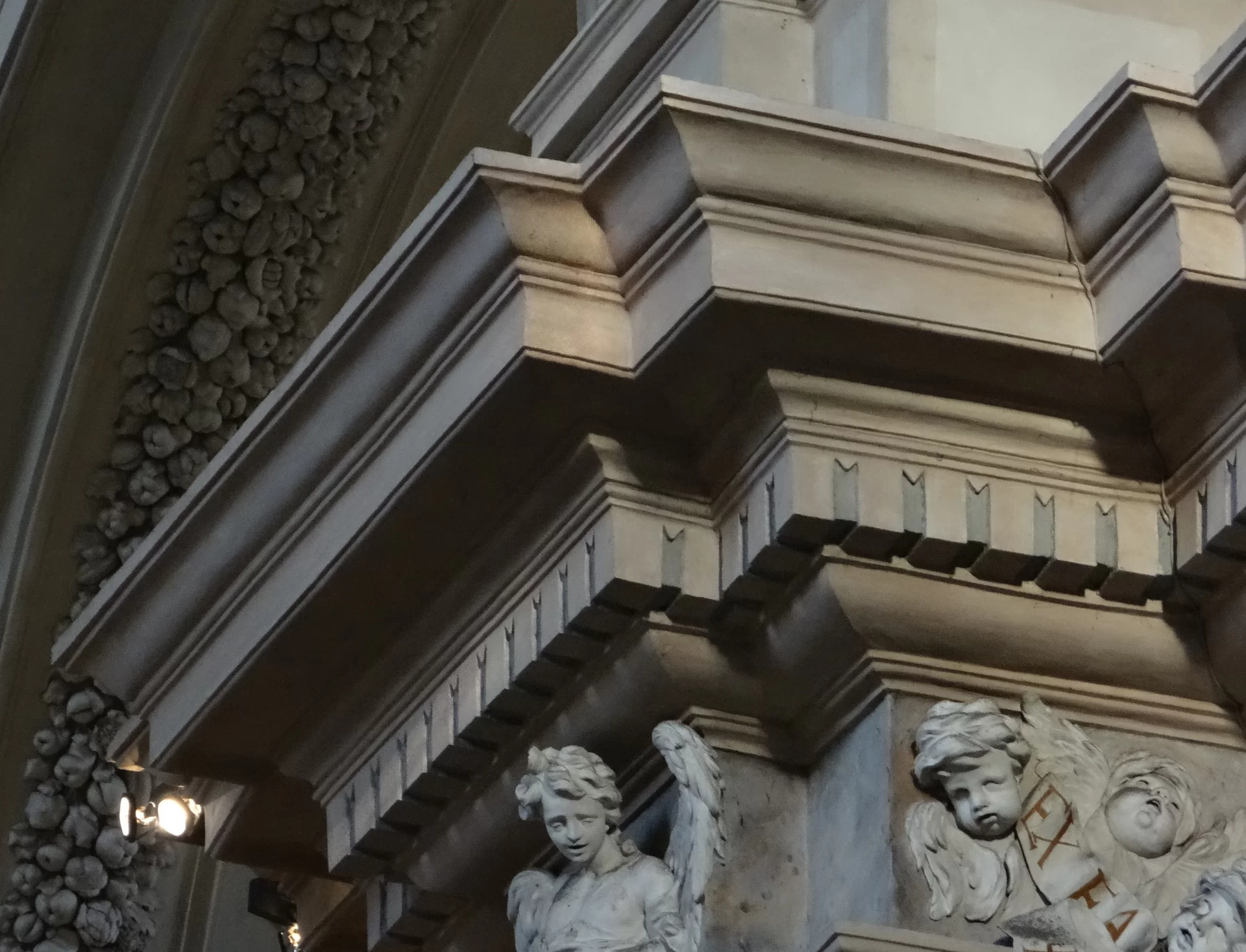 Zdjęcie przedstawia wystający poza lico ściany gzyms. Dolna część nad kolumnami posiada regularne, pionowe pasy, wyłamane ze ściany.