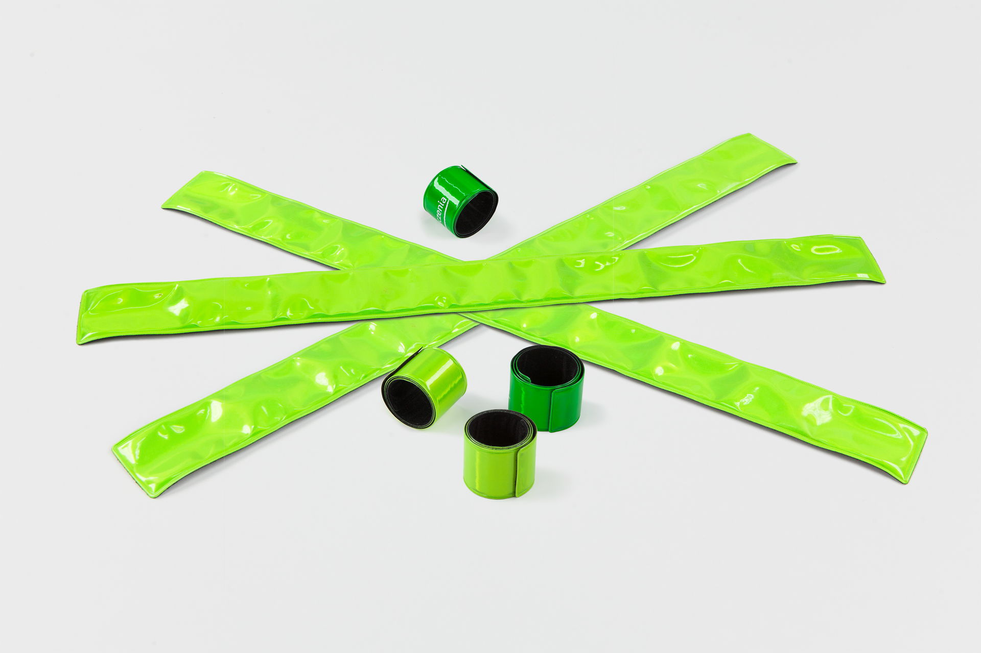 Zdjęcie przedstawia elastyczne odblaskowe zielone opaski do założenia wokół nadgarstków. Szerokość opaski to około 2 centymetry. Na zdjęciu opaski znajdują się zarówno w postaci zwiniętej jak i rozwiniętej.