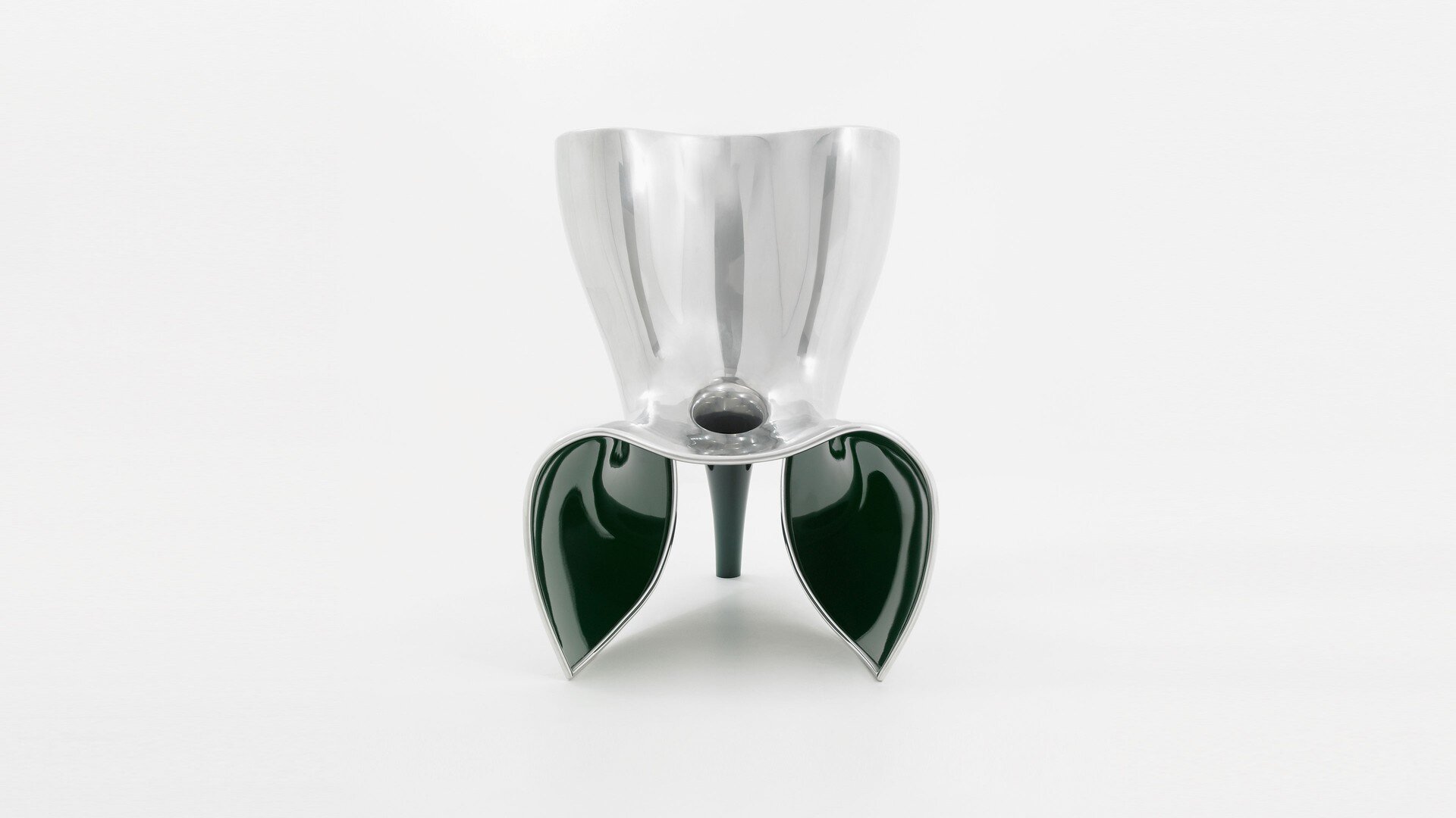 Zdjęcie przedstawia krzesło o nietypowym kształcie. Siedzisko wykonano ze srebrnego materiału o perłowym połysku. Oparcie jest prostokątne i rozszerza się ku górze. Część do siedzenia jest wygięta w dół w środkowej części, a z tyłu znajduje się przymocowany element w kształcie zwężającej się rury pustej w środku. Rura ma kolor ciemnozielony i tworzy jedną z nóg krzesła. Pozostałe dwie nogi krzesła znajdują się z przodu, po obu stronach i mają kształt płatka kwiatu. Zwężają się ku ziemi. Mają kolor ciemnozielony, ze srebrnym obramowaniem. Zdjęcie wykonano na białym tle.