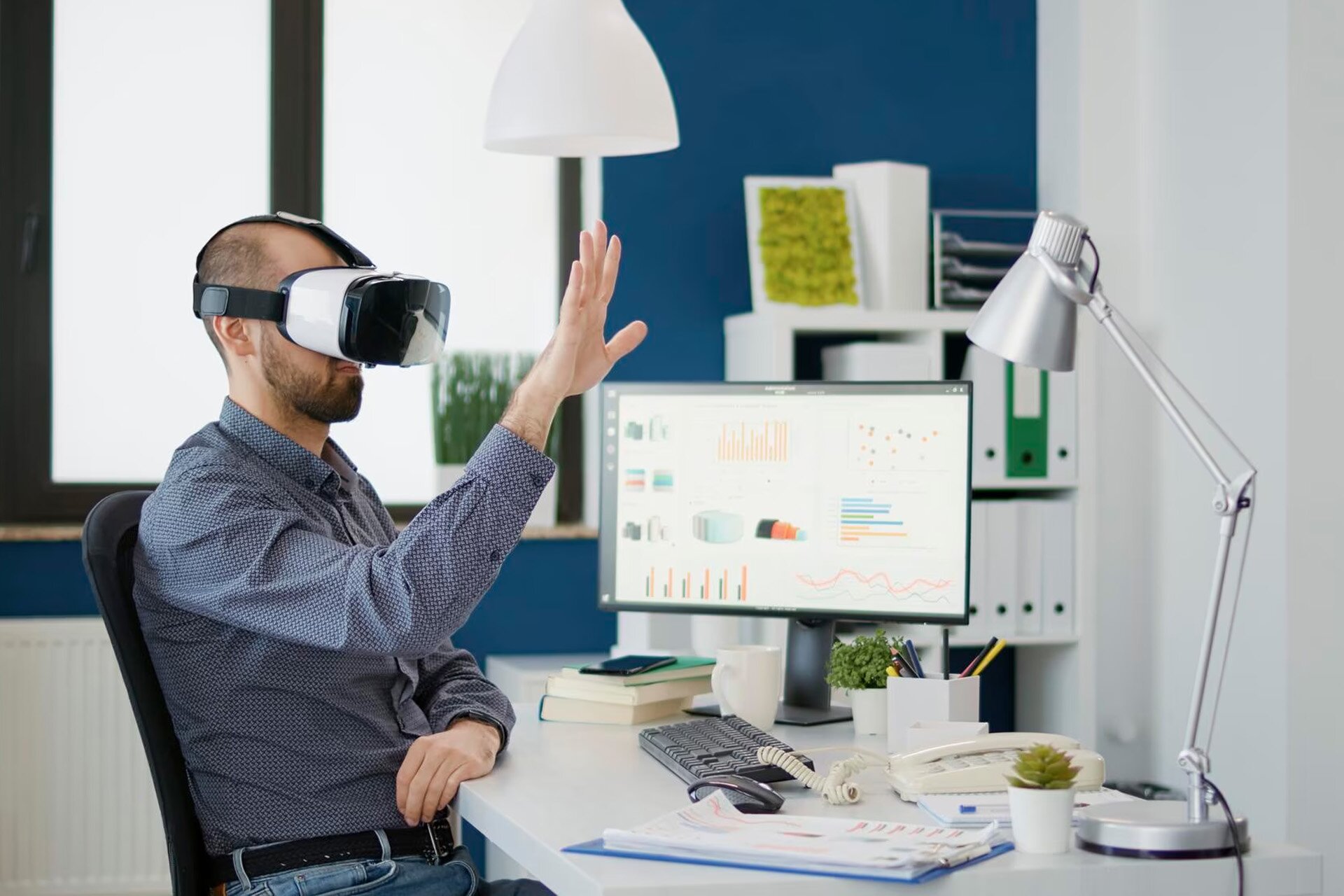 Zdjęcie przedstawia siedzącego przed monitorem mężczyznę z założonymi na głowę goglami VR. Mężczyzna  dotyka w powietrzu nie istniejącej  rzeczy. 