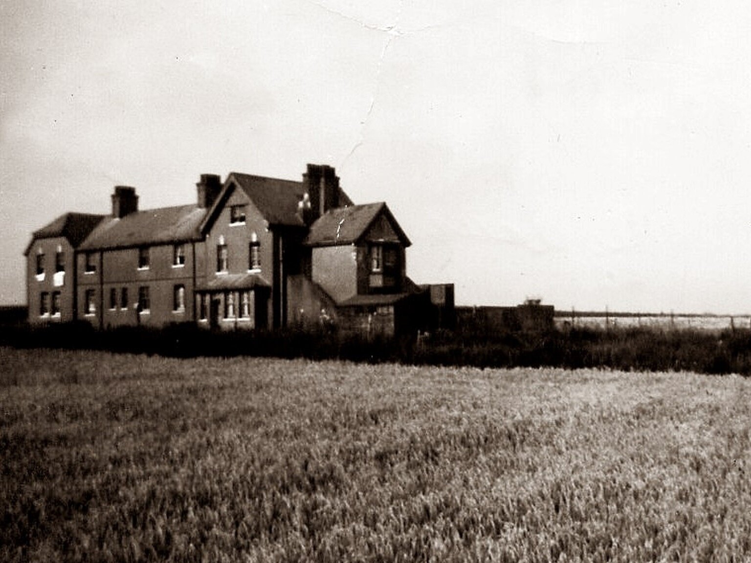 Czarno‑biała fotografia przedstawia duży dom stojący w szczerym polu. Dom składa się z kilku segmentów, ma spadzisty dach, na którym widoczne są trzy kominy.