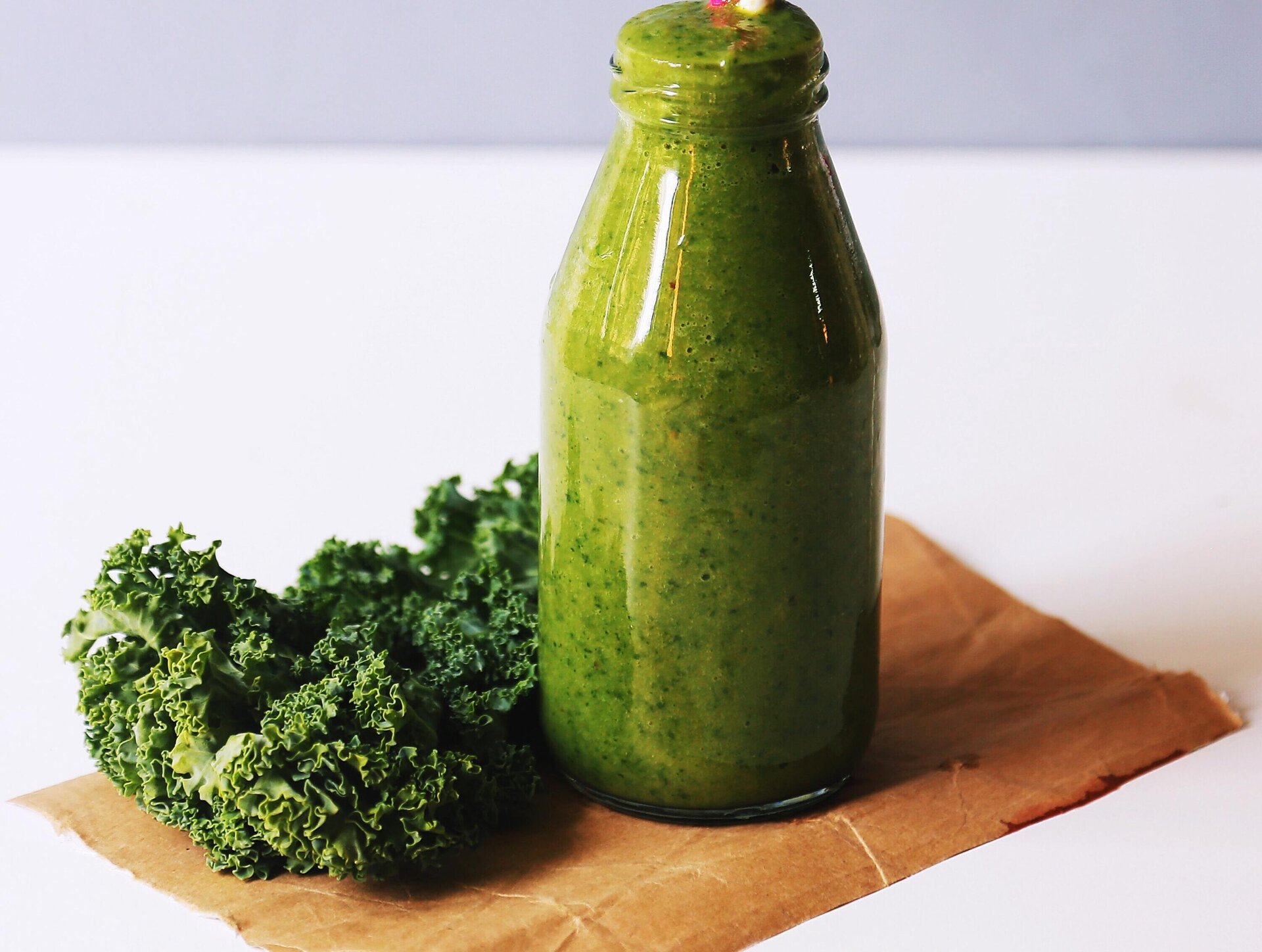 Zdjęcie przedstawia kawałek szarego papieru leżący na białym blacie. Na papierze widoczne są leżące liście jarmużu oraz szklana butelka wypełniona zielonym napojem. 