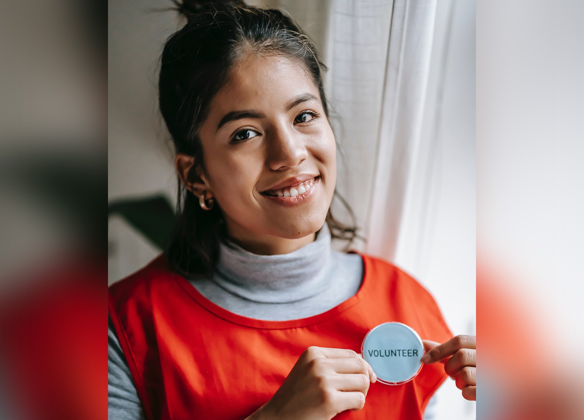 Zdjęcie przedstawia dziewczynę, w ujęciu od klatki piersiowej w górę, która w uniesionych dłoniach na wysokości klatki piersiowej trzyma okrągły znaczek z napisem w języku angielskim: VOLUNTEER. Dziewczyna jest uśmiechnięta, a na szary golf ma założoną pomarańczową kamizelkę. 