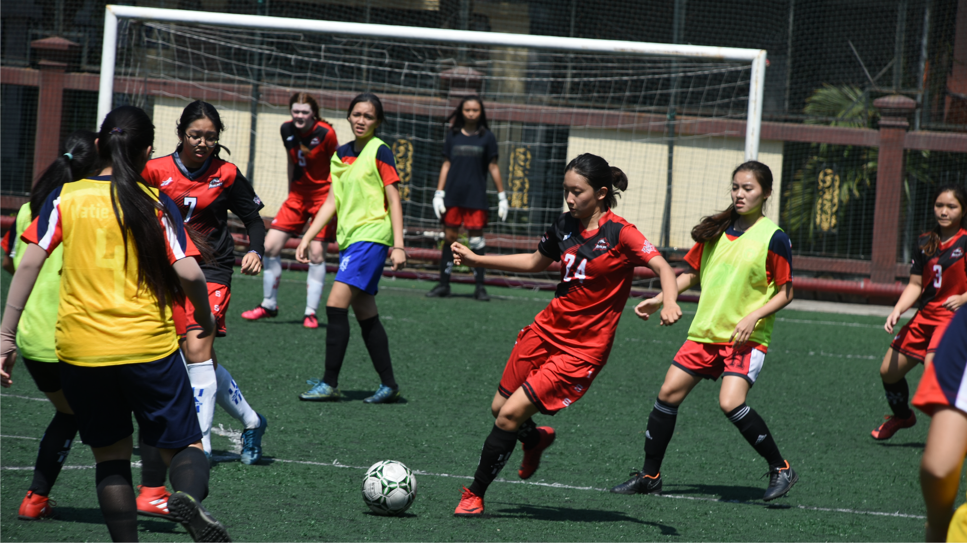 Zdjęcie przedstawia zawodniczki, dziewczyny, które grają w piłkę nożną na boisku.  Zawodniczki ubrane są w stroje do gry w piłkę nożną: koszulki, spodenki, getry i korki. Część dziewczyn na koszulki założone kamizelki odblaskowe. Zawodniczki rozgrywają piłkę w środku pola. W środkowej części zdjęcia widoczna jest dziewczyna podbiegająca do piłki i zamierza ją kopnąć. W pobliżu niej widoczne są inne zawodniczki. W tle znajduje się bramka. W bramce stoi zawodniczka w rękawicach na rękach.