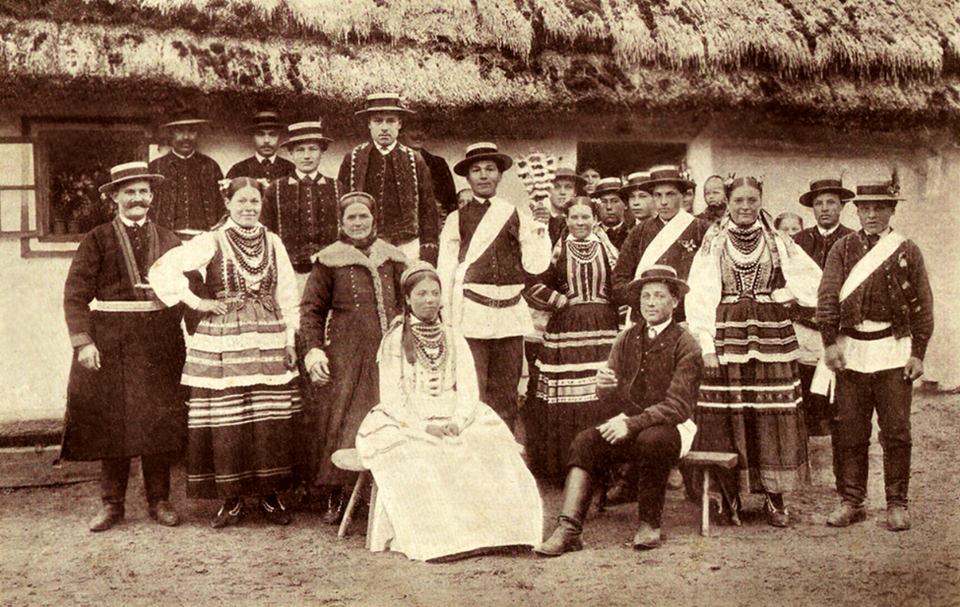 Zdjęcie przedstawia gości weselnych i młodą parę pozujących do zdjęcia po ślubie. Wszyscy zebrani są w ludowych strojach. Za ich plecami znajduje się chałupa kryta strzechą.