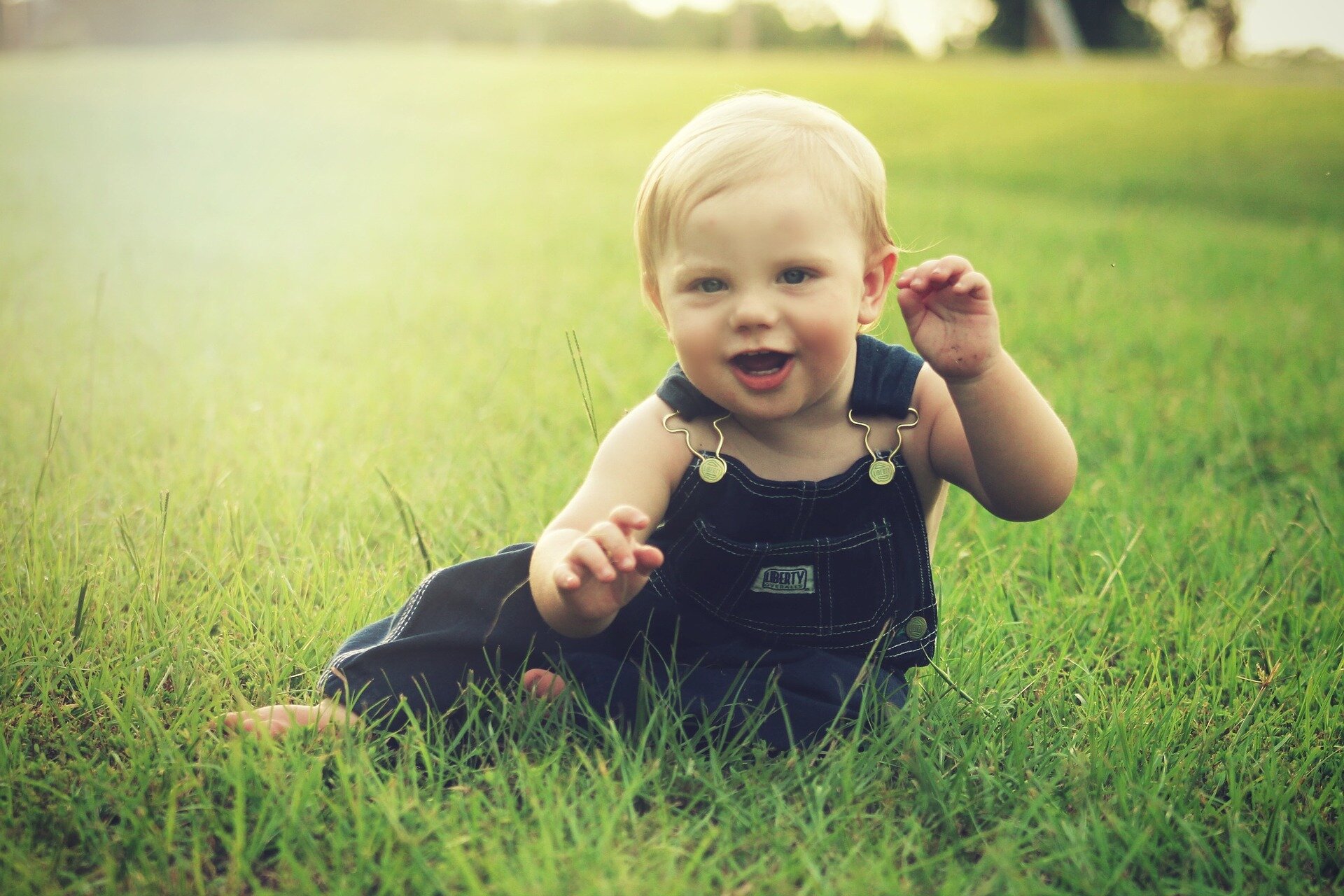 Zdjęcie przedstawia małego chłopca w ogrodniczkach siedzącego na trawie. Chłopiec uśmiecha się.