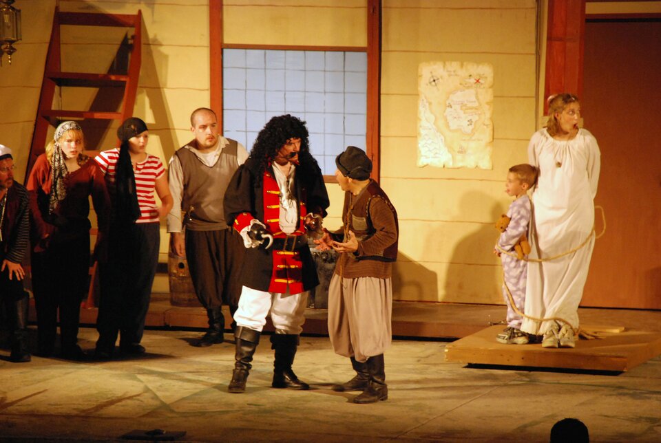 Ilustracja przedstawia scenę z teatru. W centrum zdjęcia widać dwóch rozmawiających mężczyzn, którzy są aktorami. Dookoła stoją inni ludzie, również aktorzy. Z lewej strony pomieszczenia do ściany doczepiona jest czerwona drabina. Postaci są poprzebierane w stroje pirackie, z wyjątkiem dwóch osób z prawej strony - kobieta i dziecko stoją w piżamach i są do siebie przywiązani sznurkiem.