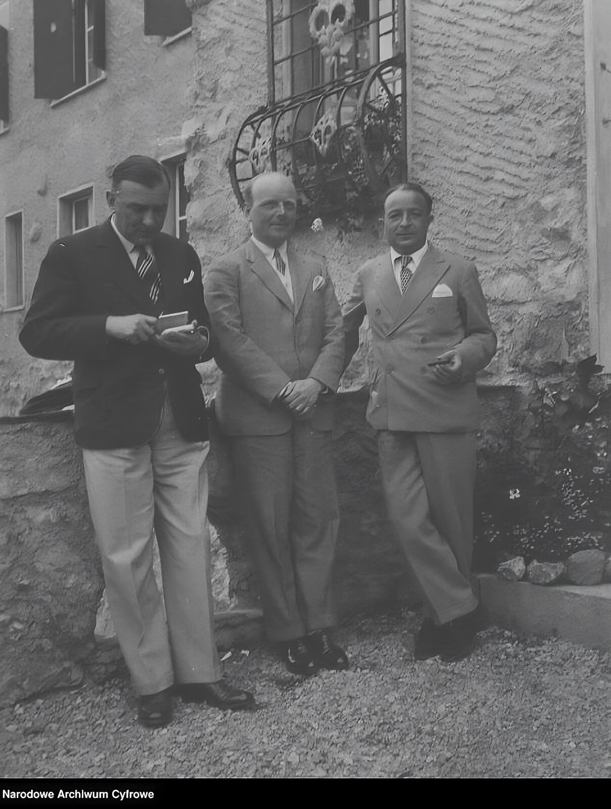 Zdjęcie przedstawia trzech mężczyzn, stojących przy murku kamiennej budowli z okratowanym balkonem. Dwaj noszą szare garnitury, krawaty poszetki w kieszonkach marynarek.  Stojący po lewej stronie Szymanowski ma marynarkę ciemną marynarkę z poszetką w kieszeni, krawat oraz jasne spodnie. Stojący w środku hrabia Potocki założył przed sobą dłonie.  Kochański ma prawą dłoń opartą na murku, a lewą z cygarem zgiętą przed sobą. Po prawej stronie zdjęcia widoczny jest skalniak z roślinnością ogrodową. 