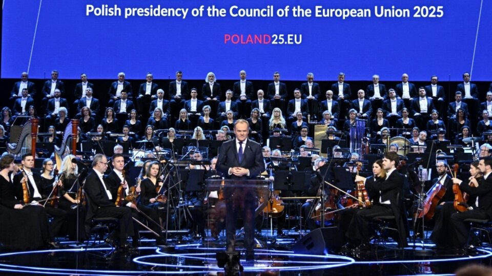 Premier Polski stoi na scenie przed dużą orkiestrą i chórem, przemawiając podczas inauguracji polskiej prezydencji w Radzie Unii Europejskiej 2025. Na ekranie za nim widnieje napis: ‘Polish presidency of the Council of the European Union 2025, POLAND25.EU’.