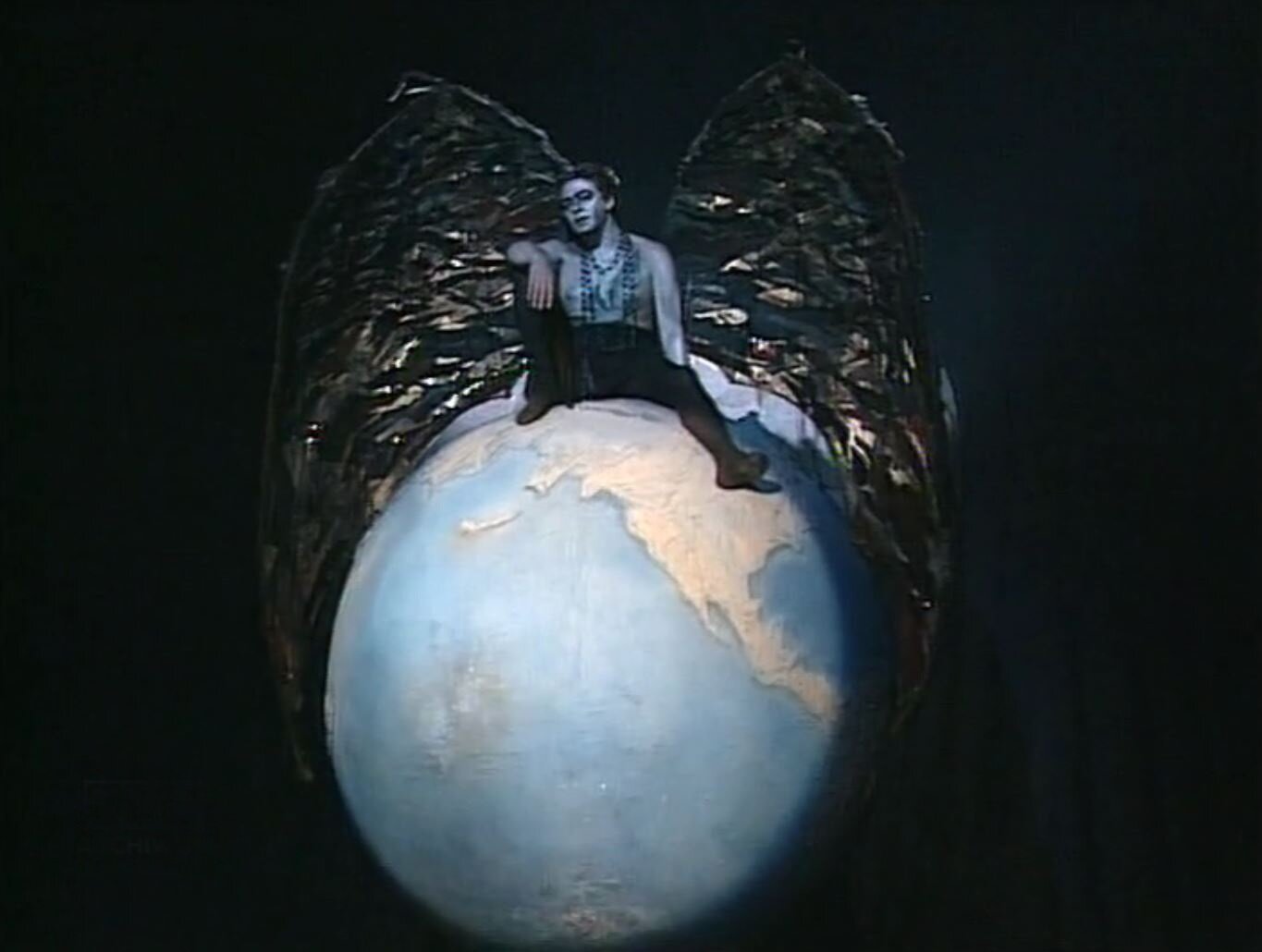 Zdjęcie przedstawia kadr z filmu. To zbliżenie na dużą kulę ziemską. Siedzi na niej szatan. Ma on postać dojrzałego mężczyzny z ogromnymi skrzydłami. Tors szatana jest nagi, zawieszony jest na nim długi sznur korali.