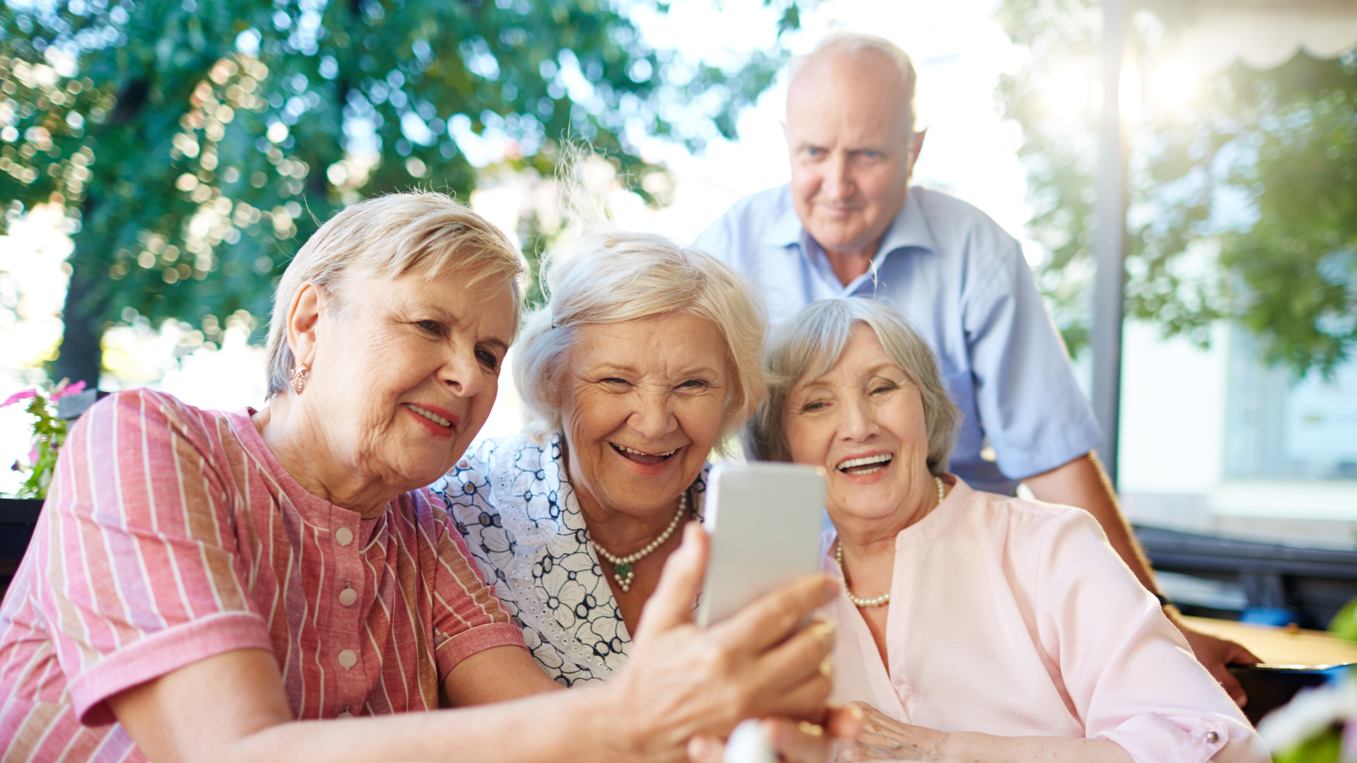 Zdjęcie przedstawia trzy starsze kobiety oraz stojącego za nimi starszego mężczyznę. Kobiety siedzą, są uśmiechnięte. Kobieta po lewej stronie na zdjęciu trzyma w ręku telefon komórkowy i pokazuje coś innym na ekranie lub wykonuje zdjęcie typu selfie. Zdjęcie zostało wykonane na świeżym powietrzu, w tle widoczne są drzewa.