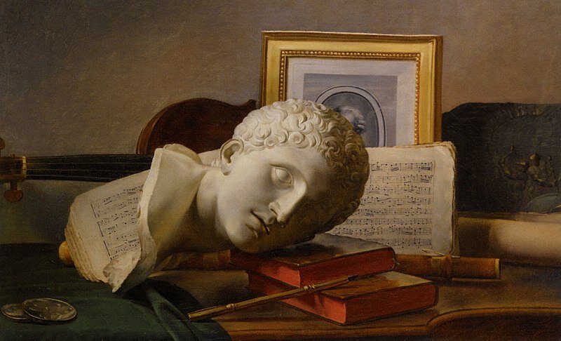 Obraz przedstawia mały posążek popiersia mężczyzny, który leży na biurku. Na biurku znajdują się książki, pióro, otwarty zeszyt nutowy, skrzypce oraz mały portret.