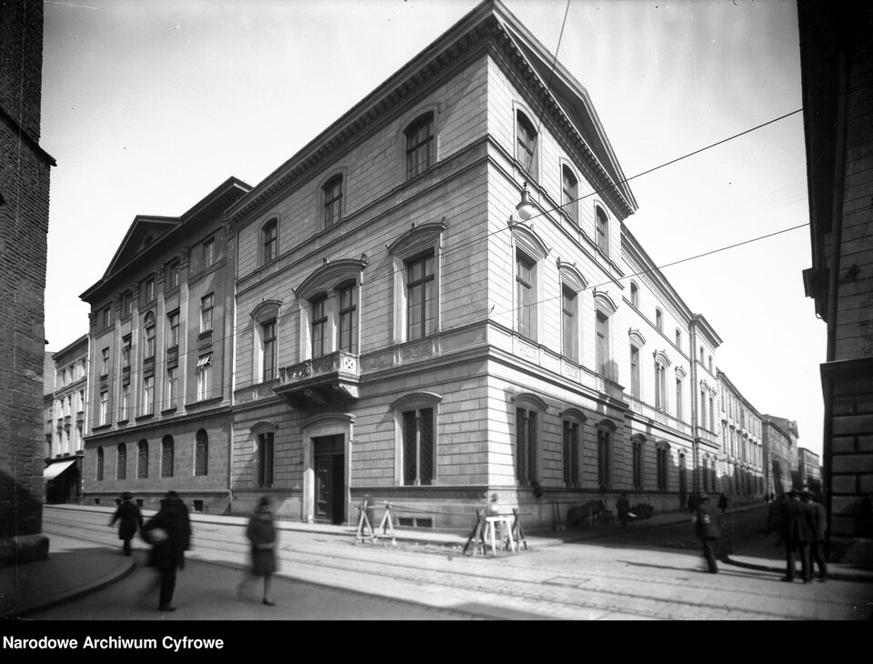 Na zdjęciu dwupiętrowy budynek. Stoi na rogu ulic. Po chodniku idą ludzie.