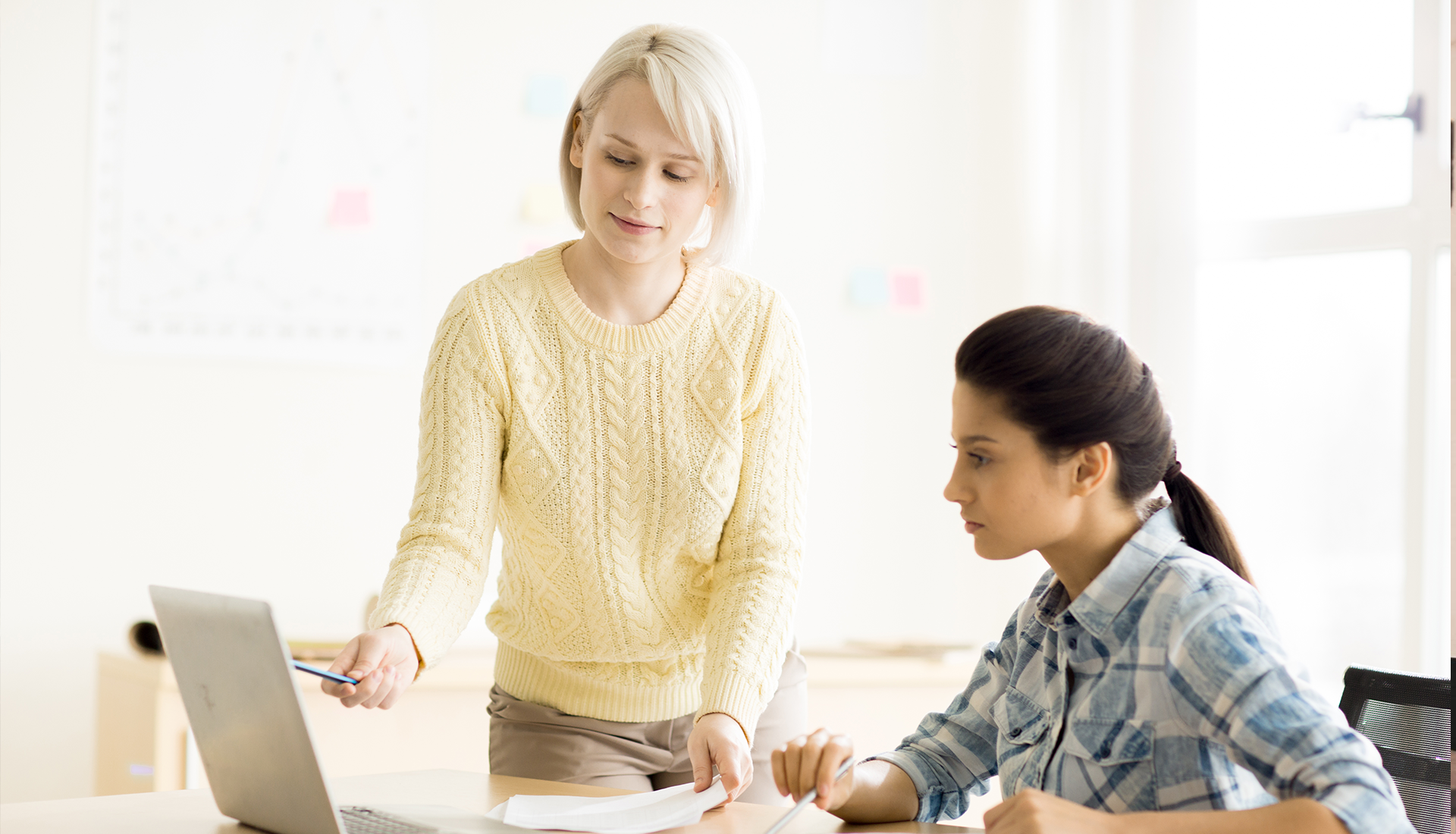Zdjęcie przedstawia dwie młode kobiety, w ujęciu od pasa w górę. Kobieta po prawej, widoczna z lewego boku, siedzi przy biurku. Ma długie, ciemne włosy splecione z tyłu. Ubrana jest w kraciastą koszulę. Obie ręce ma położone na blacie, a w prawej ręce trzyma długopis. Patrzy na monitor otwartego laptopa stojącego przed nią na biurku. Kobieta po jej lewej stronie stoi za biurkiem. Ma krótkie blond włosy i jest ubrana w jasnobrązowe spodnie oraz żółty sweter. W prawej ręce trzyma długopis i wskazuje coś na ekranie laptopa, zaś w lewej ręce trzyma kartki, które kładzie na blacie. W tle, po prawej, widoczne jest duże okno.