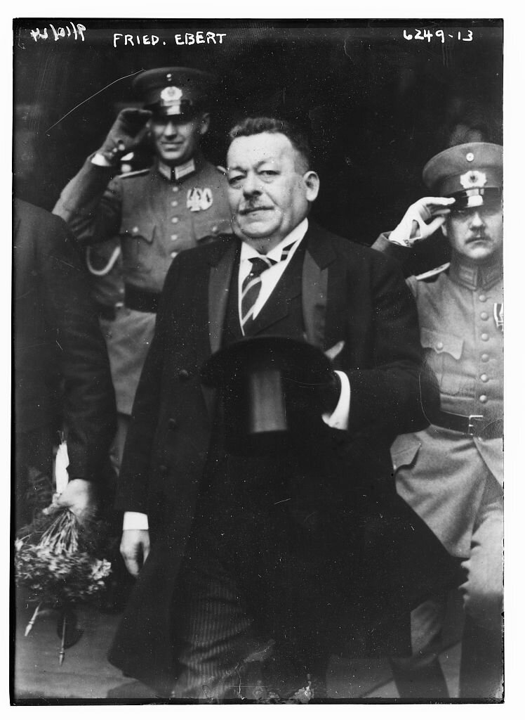 Kliknij, aby powiększyć Zdjęcie przedstawia trzech mężczyzn. Wśród nich obecny jest korpulentny prezydent Friedrich Ebert. Ubrany jest w czarny surdut, ma białą koszulę i krawat. Pod nosem ma wąskie wąsy. Jego włosy są krótkie, ma widoczne zakola. W ręku trzyma cylinder. Pozostali dwaj mężczyźni stoją za prezydentem. Ubrani są w mundury. Na głowie mają czapki wojskowe. Prawą ręką salutują. U góry na fotografii dopisany jest napis Fried. Ebert.
