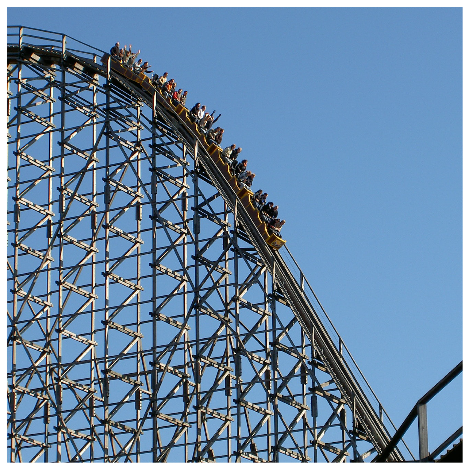 Na zdjęciu widoczna jest kolejka górska. W wagonikach zjeżdżających po stromej górce siedzą ludzie. W tle widoczne jest błękitne niebo.  