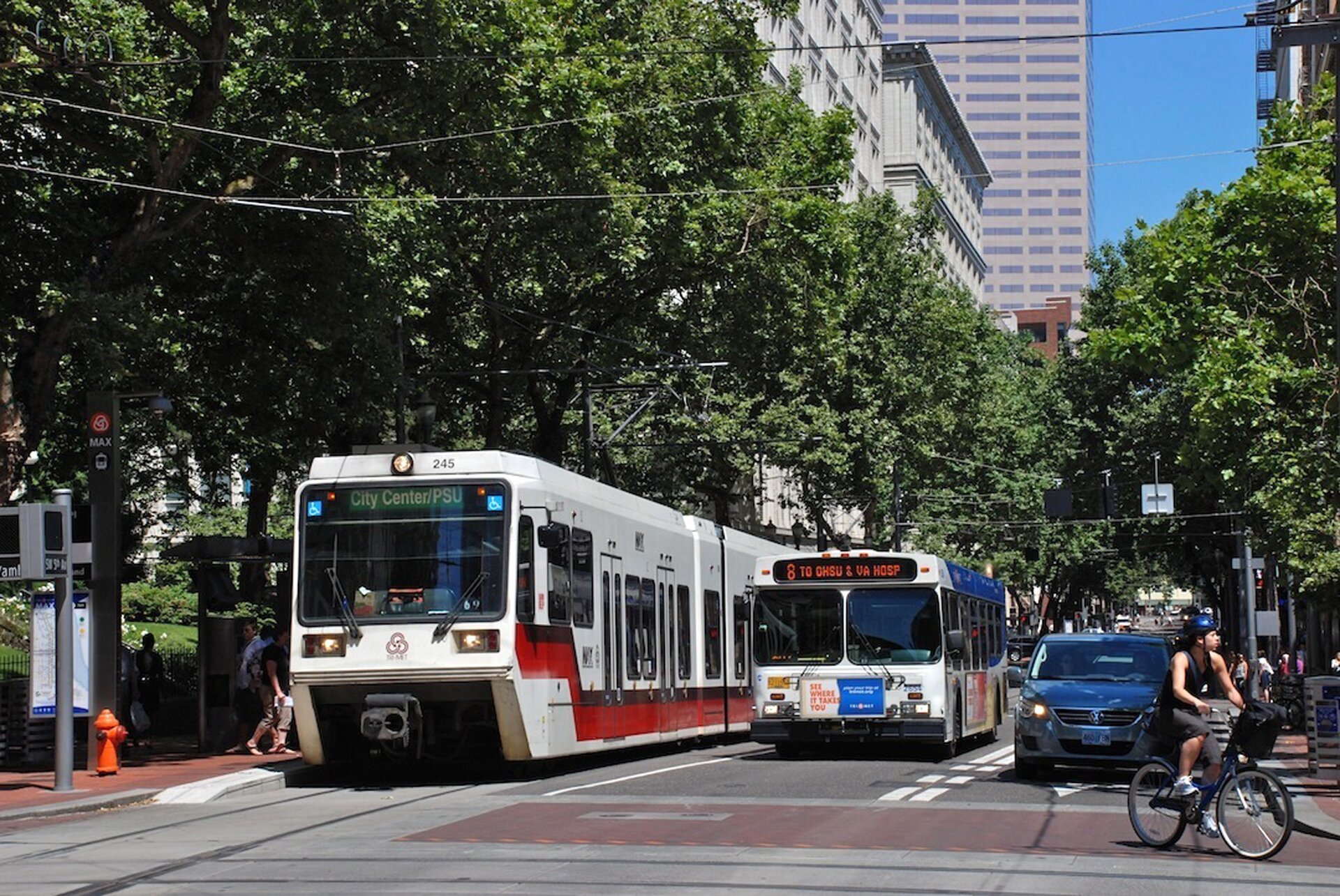 Fotografia przedstawia ulicę w Portland. Słoneczny dzień, jadący rowerzysta, tramwaj, autobus, zielone drzewa.