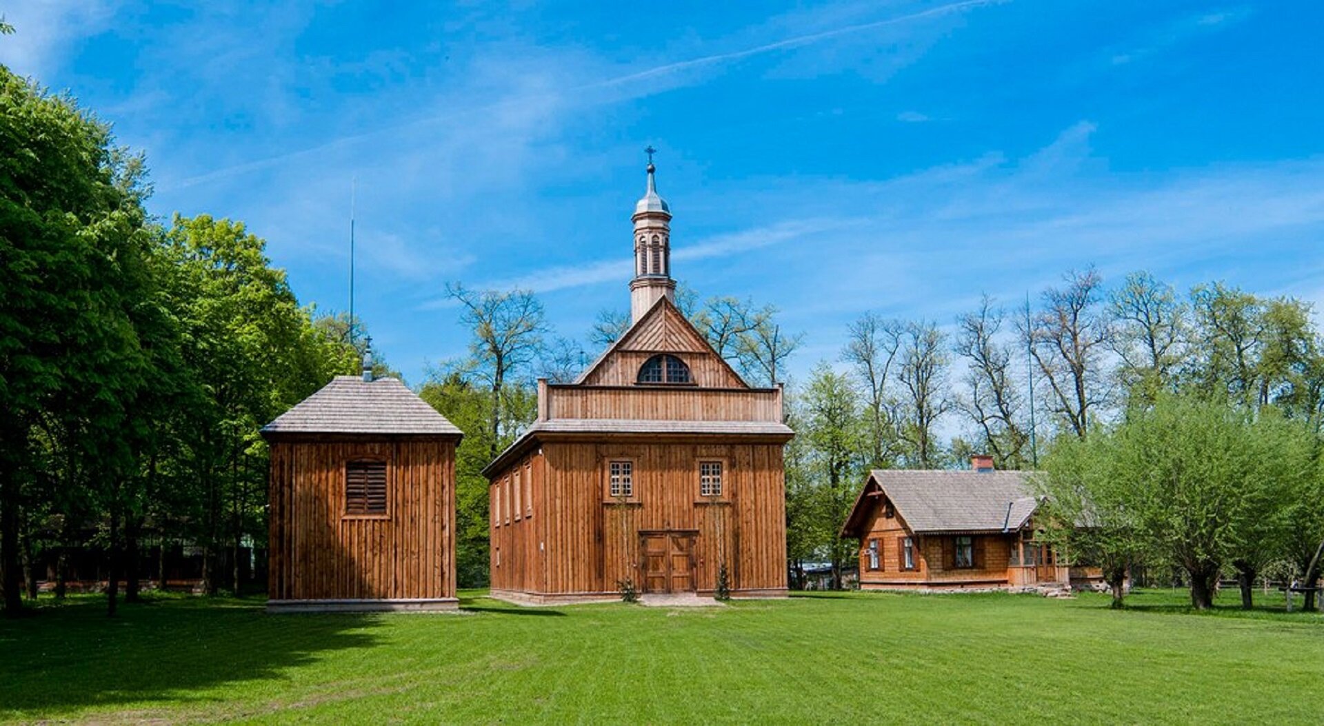 Zdjęcie ukazuje drewniany kościół. Budynek jest brązowy. Obok głównego budynku stoją dwa mniejsze drewniane budynki. Kościół, ma dwa centralnie umieszczone okna oraz drzwi. Na dachu budynku znajduje się, najprawdopodobniej dzwonnica. Przed budynkiem rozciąga się zielona trawa, a w tle ukazane jest błękitne niebo.