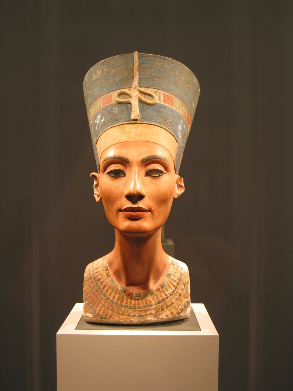 Fotografia przedstawia Popiersie Nefertiti (Nefretete). Ukazuje kobietę, której głowa jest osadzona na długiej szyi. Jej ciemnoniebieskie i podkreślone mocnym konturem oczy są lekko przysłonięte powiekami. Na jej ustach rysuje się lekki uśmiech. Kobieta ma wyeksponowane kości policzkowe subtelny policzki, a na powiekach, poniżej oczu oraz obok nosa widoczne są fałdy skórne. Na skroniach Nefertiti spoczywa wysoki ciemno‑błękitna tiara zdobiona złotym diademem królewskim z prostokątnymi elementami w zielonkawych i rdzawych tonacjach. Z przodu diademu znajduje się ozdoba w postaci węża.