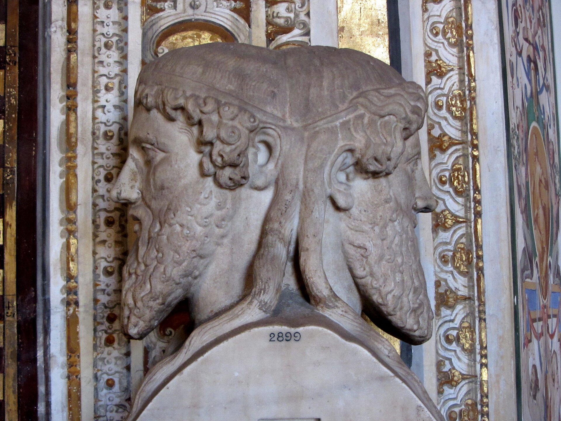 Na zdjęciu przedstawiona została rzeźba w jasnym kolorze, ukazująca dwóch mężczyzn o kręconych włosach oraz brodach. Mężczyźni stoją do siebie plecami; wydaje się że są ze sobą scaleni. Tuż pod szyjami widnieją cyfry 2899.