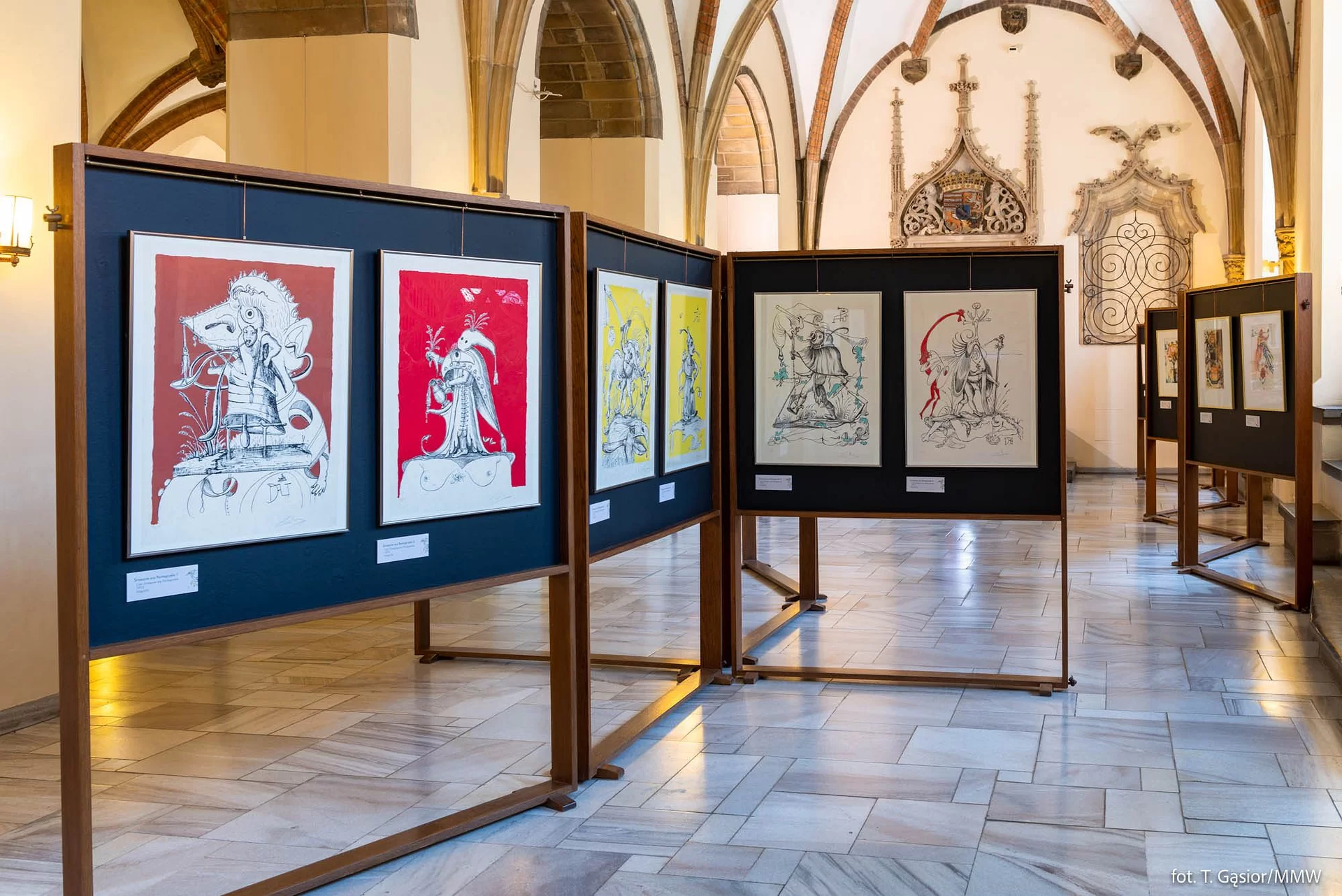 Zdjęcie przedstawia fragment wystawy. Prace artysty zaprezentowano na drewnianych tablicach – na każdej z tablic umieszczono po dwa dzieła artysty. Wnętrze, w którym znajdują się prace posiada kwadratowe kolumny, a miedzy nimi łukowe przejścia. Ściany są jasnobeżowe.