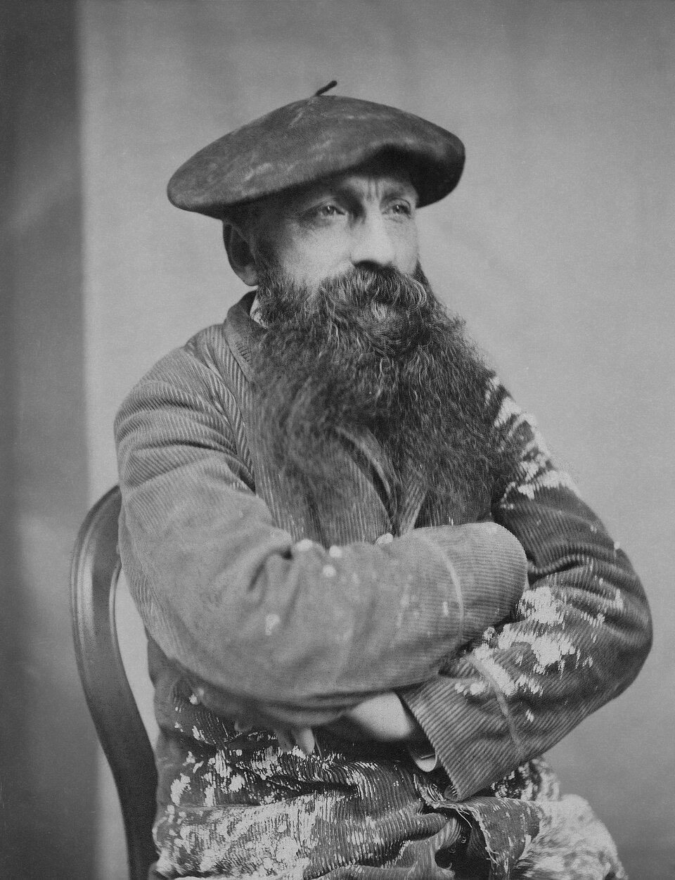 Czarno‑biały portret Auguste’a Rodina siedzącego na krześle. Artysta ma na głowie szeroki beret, długą, gęstą brodę i wąsy. Ubrany jest w ciężką, prążkowaną marynarkę lub bluzę. Ma skrzyżowane na piersi ręce i patrzy w bok, poza kadr. W tle znajduje się jednolite, jasne tło studyjne.